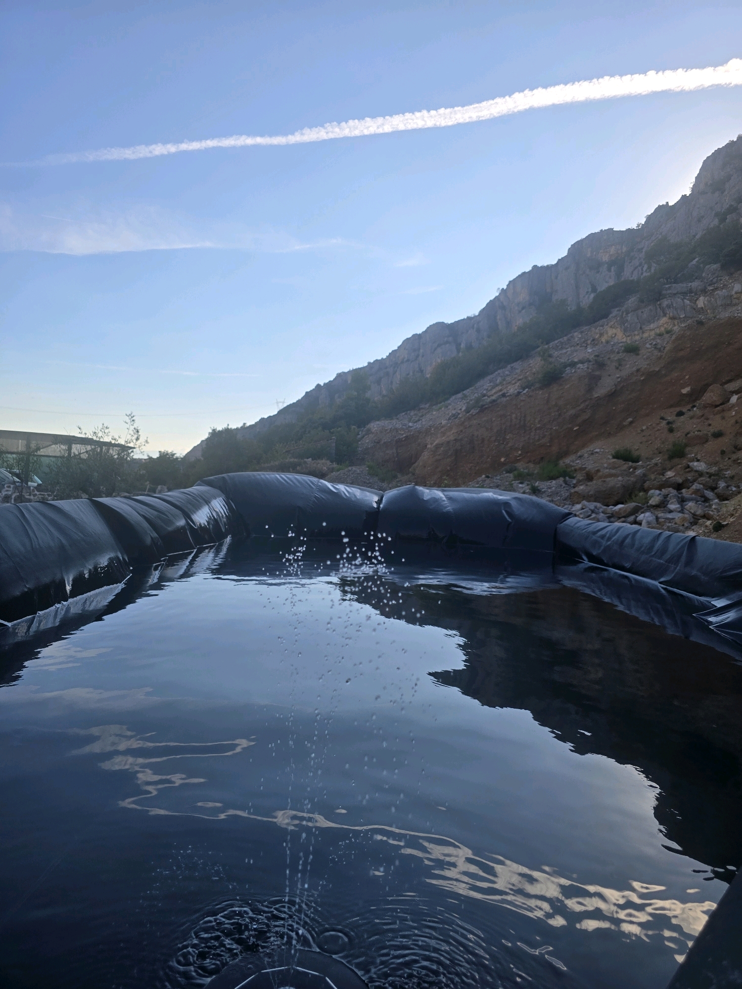 This media file depicts an outdoor scene featuring a newly installed pond or water reservoir lined with black material. The water within the pond is dark and reflects the sky and surrounding landscape. Small jets of water are visible, creating a spray and ripples on the surface. The pond is situated in a sloped, rocky, and arid environment, characteristic of a Mediterranean climate. In the background, a steep, rocky mountainside rises, with sparse vegetation clinging to its slopes. The sky is a clear blue with a thin, elongated white contrail streaking across it, suggesting it is daytime. The lighting indicates it may be late afternoon or early morning, with the sun casting a gentle glow on the mountain. Structures, possibly associated with construction or agricultural activity, are visible in the far distance on the left. The overall impression is one of a newly created water feature in a rugged, natural setting, possibly for irrigation or agricultural purposes. The location context is Podgorica, Montenegro.