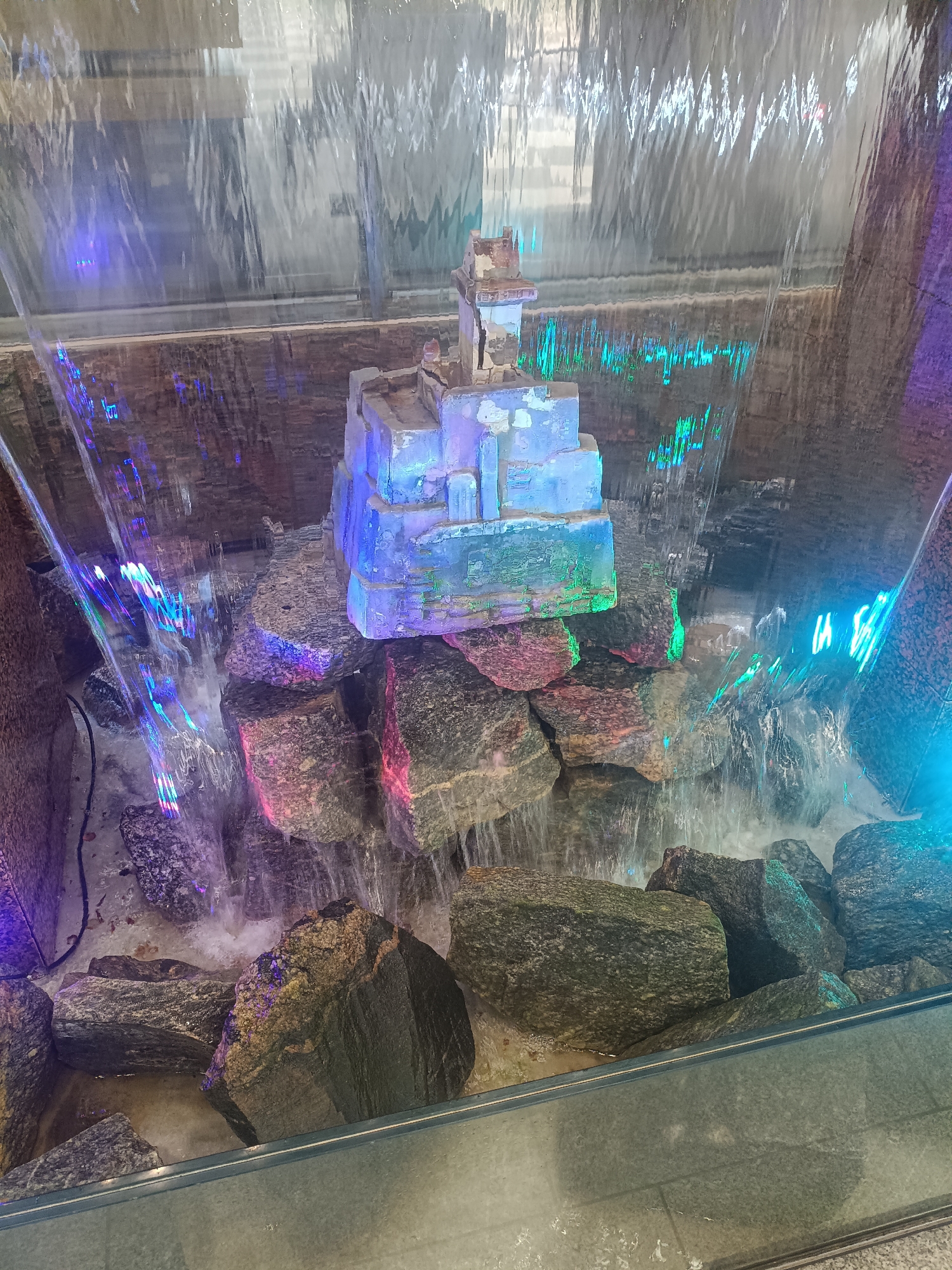 This image shows a close-up of an indoor water feature with artificial rock formations and a cascading water curtain. The water is illuminated by colored lights, predominantly blue and green, creating a vibrant and somewhat surreal atmosphere. There is a small, multi-tiered structure resembling a miniature castle or building placed on top of some of the rocks. The rocks themselves are large and irregular, some with pinkish or reddish hues, others dark grey or black. Water flows over and around these rocks, creating a sense of movement and sound. The background is blurred but appears to be a wall with a textured surface, possibly stone or brick, also lit by colored lights. The foreground shows a clear barrier, likely glass, through which the water feature is viewed. The overall impression is one of a decorative, possibly commercial or public space installation. The image does not provide any visual cues to indicate its location in Narva, Estonia.