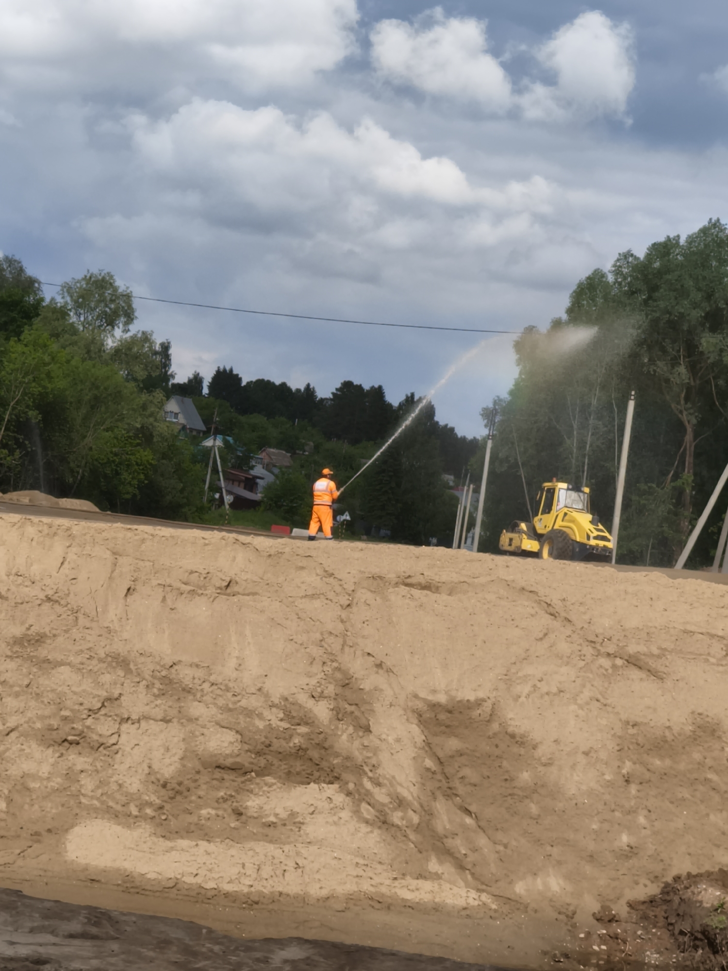 The image depicts an outdoor construction site in Oktyabr'skii, Russia, on an overcast day. In the foreground and mid-ground, a large, uneven embankment of light brown sand or dirt stretches across the frame.
On top of this elevated ground, a single construction worker is visible. Dressed in bright orange protective work overalls and a hard hat, the worker is standing with their back mostly towards the camera, actively operating a long hose. A powerful stream of water emanates from the hose, arcing through the air and landing on the sandy surface, likely for dust suppression or soil compaction. A faint rainbow is visible within the water spray, suggesting sunlight refracting through the droplets despite the cloudy sky.
Further to the right on the same level, a bright yellow construction vehicle, appearing to be a soil compactor or road roller, is parked, indicating heavy machinery is involved in the ground preparation.
The background features a verdant landscape with numerous green trees and several utility poles. Beyond the trees, glimpses of residential houses with pitched roofs are discernible, suggesting the construction is taking place in a suburban or semi-rural area. The sky is largely covered with a mix of grey and white clouds. The scene captures an active phase of infrastructure development or land preparation. No visible text is present.