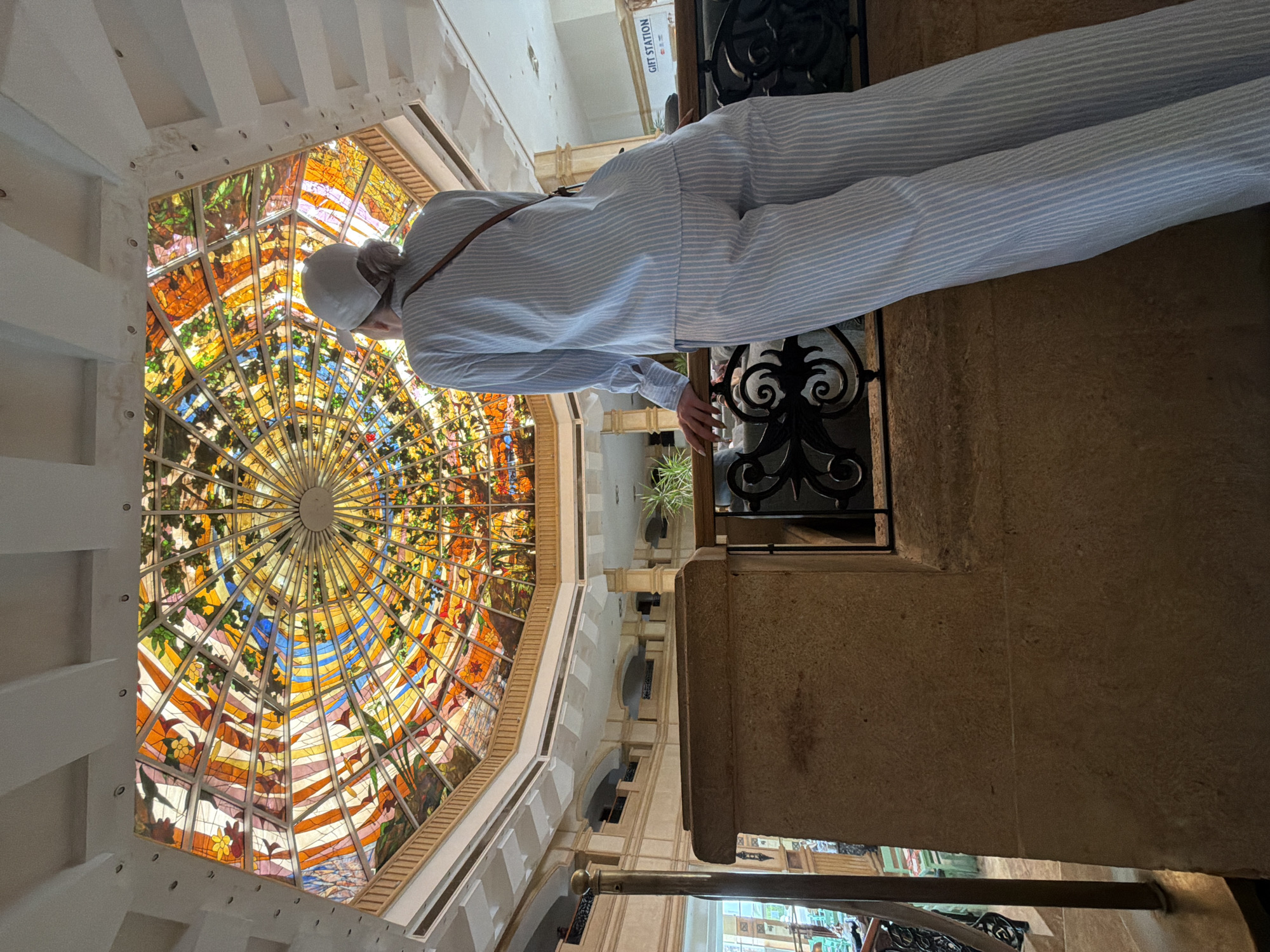 A medium shot shows a person standing in an interior space with a large, ornate, stained-glass ceiling. The person is facing the stained-glass ceiling, which is the focal point of the image. The person is wearing a white baseball cap and a light-blue-and-white striped shirt with matching pants. They lean their hands on a black, ornate railing. The background reveals an open space with multiple levels and archways, suggesting the interior of a grand building. A sign reading "GIFT STATION" can be seen in the background, likely indicating the presence of a souvenir shop. The image is captured at a slightly tilted angle, enhancing the perspective of the high ceiling and the architectural details of the building. Considering the architectural style, it is likely this image was taken at the Al-Mustafa Mosque, located in Sharm El Sheikh, Egypt.