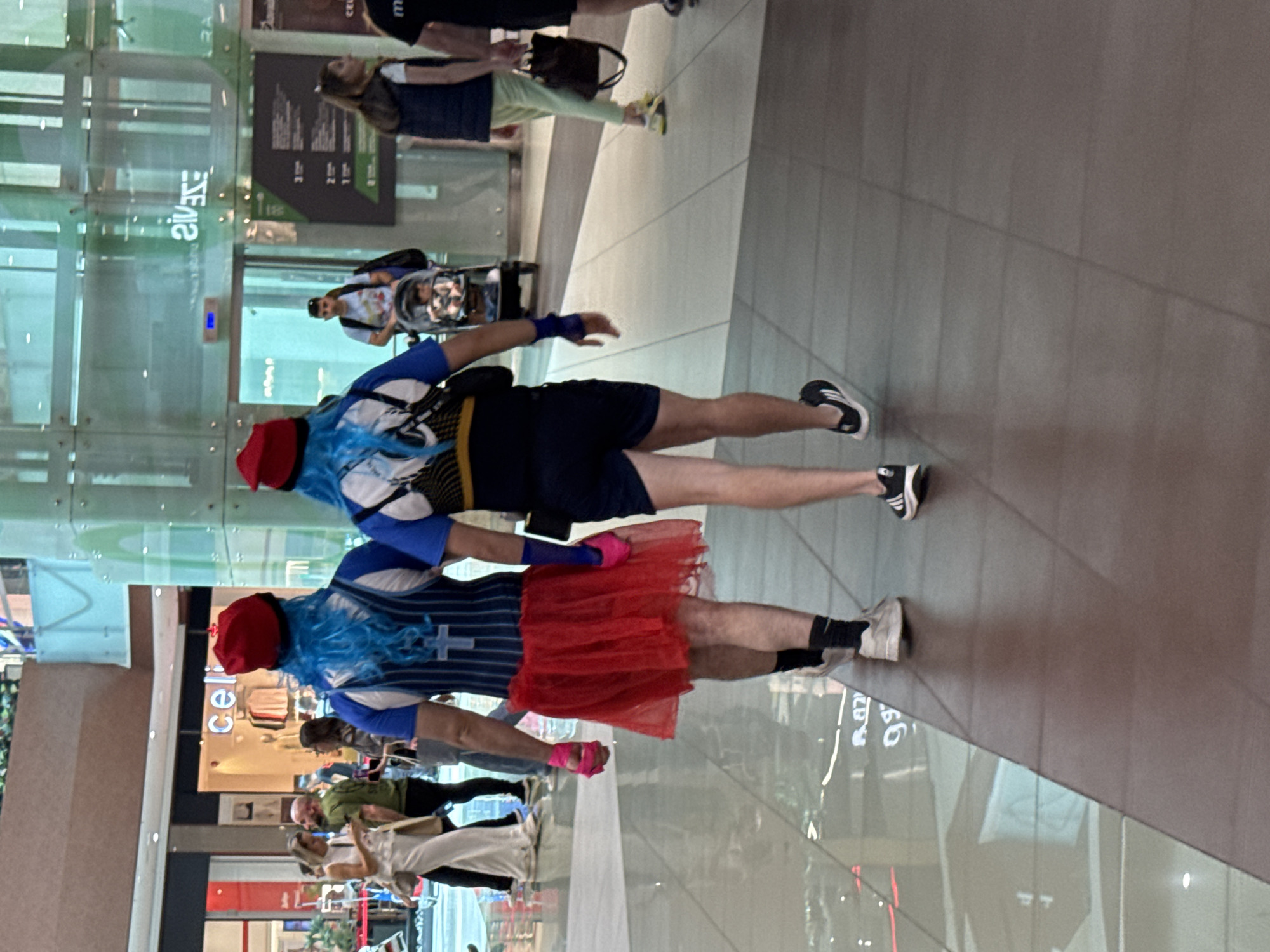 The photograph depicts two individuals, potentially associated with a running event, in what appears to be a shopping mall. The location is likely Sofia, Bulgaria based on visual clues (but I can't confirm with certainty).
The first individual is wearing a blue wig, a red beanie, a blue and white running top with a cross symbol, a red tutu, and pink wristbands. They are wearing sneakers with black socks.
The second individual is wearing a blue wig, a red beanie, and a dark running outfit.
Both individuals are walking side by side through the mall.
The mall environment includes glass walls, other shoppers, storefronts, and tiled flooring.