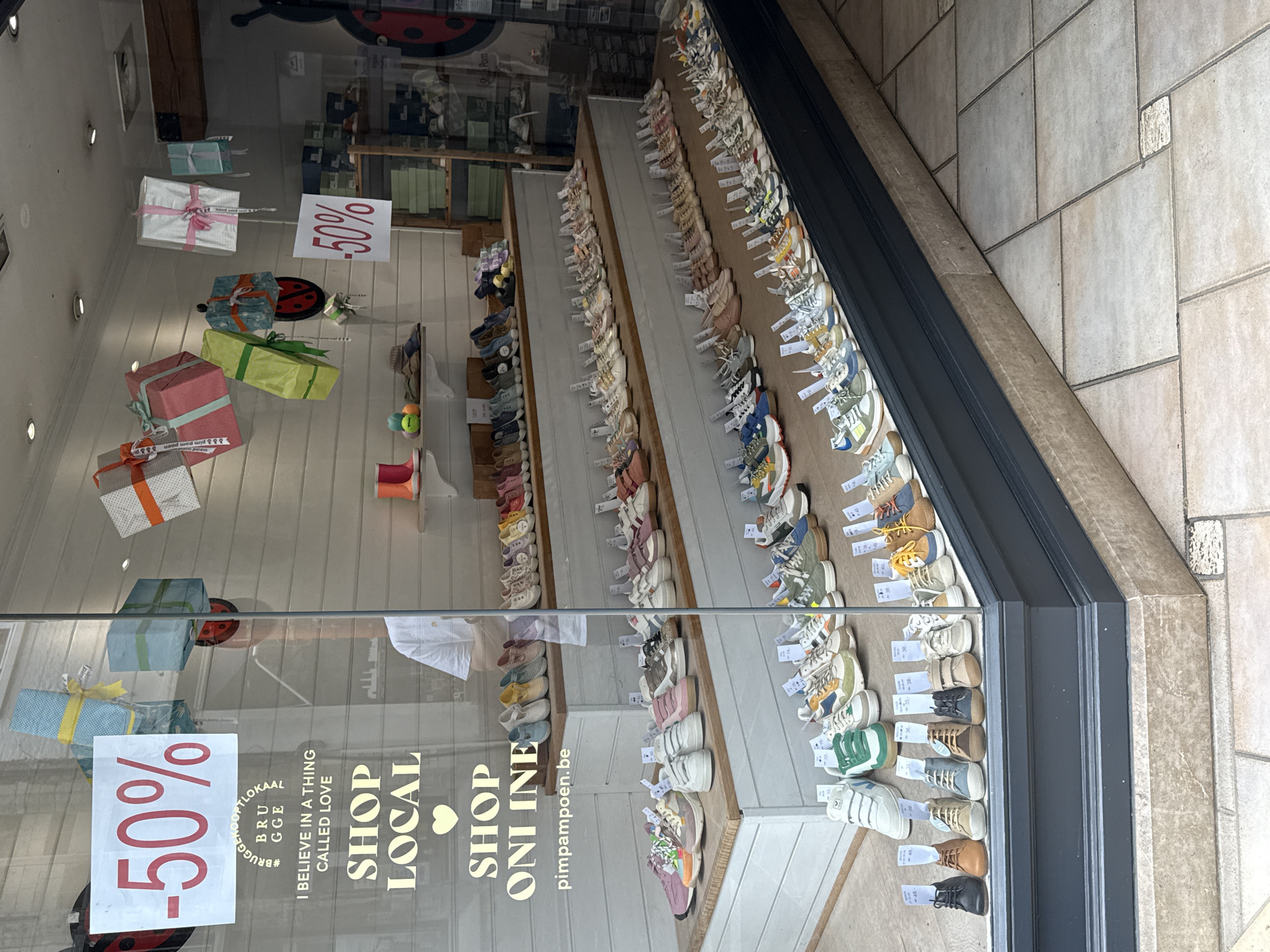 The image shows the storefront window of a shoe store located in Bruges, Belgium. A large sign with "-50%" is prominently displayed on the left side of the window. Below this sign, text reads "#BRUGGEKOPTLOKAAL BRUGGE I BELIEVE IN A THING CALLED LOVE SHOP LOCAL SHOP ONLINE pimpampoen.be". The window displays various shoes, including sneakers and children's shoes, arranged on shelves and the floor. Several gift-wrapped boxes are suspended from the ceiling of the window display, some decorated with ladybug motifs. In the background, a person wearing a white t-shirt is visible inside the store, appearing to be an employee. The flooring outside the store is made of light-colored square tiles.