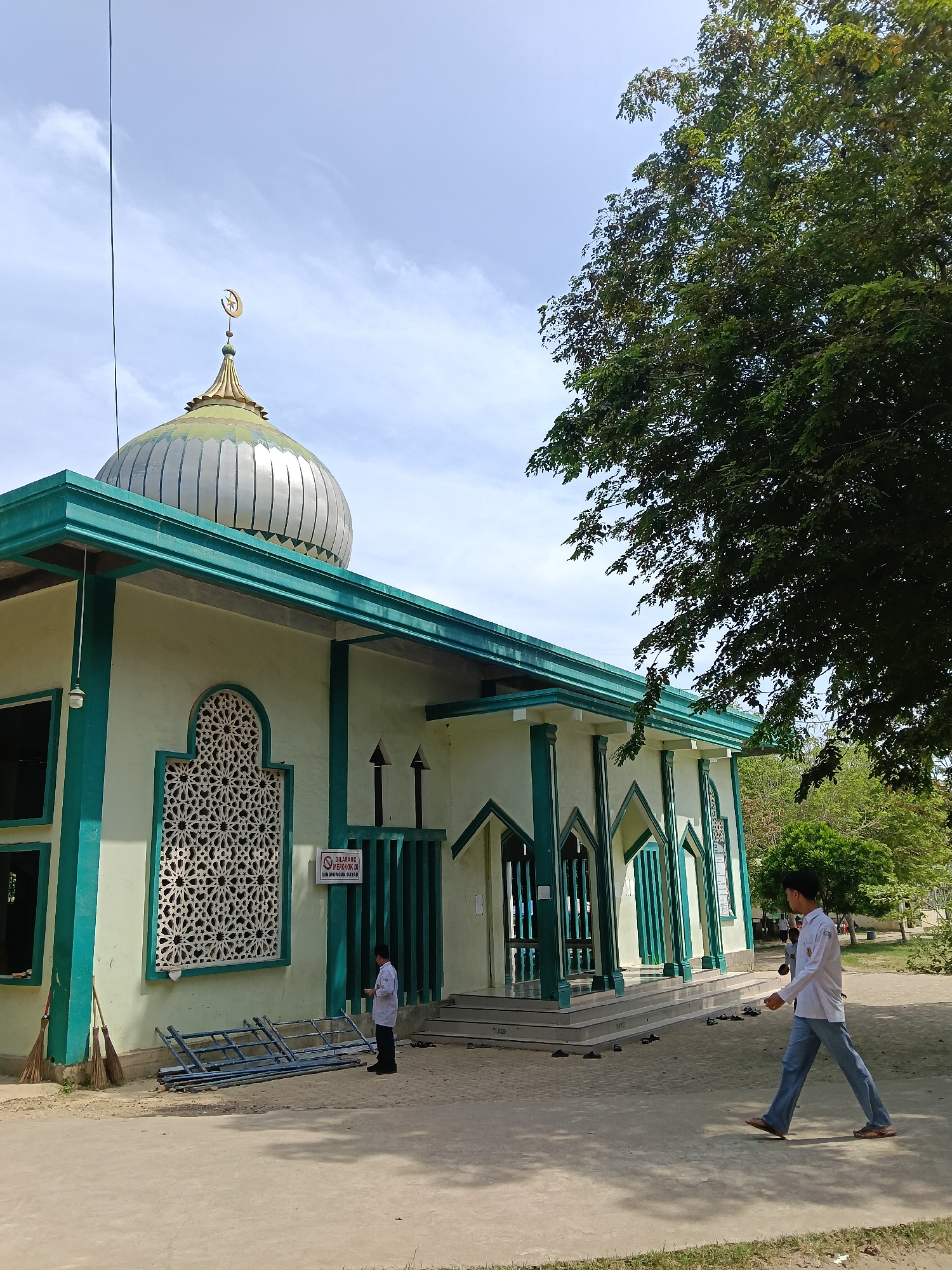 This image captures the exterior of a mosque on a bright, sunny day in Lhokseumawe, Indonesia. The building features light cream-colored walls contrasted with prominent teal or dark green trim, pillars, and roof edges. A large, silver-ribbed dome, topped with a golden crescent moon and star finial, crowns the mosque, set against a clear blue sky with some scattered white clouds.
An intricate Islamic geometric pattern adorns a recessed window or vent on the left side of the mosque's facade. Several pairs of sandals and shoes are neatly placed on the ground near the entrance steps, suggesting worshippers have entered the building.
Two individuals are visible. In the foreground, a man wearing a white long-sleeved shirt, blue jeans, and sandals walks across a concrete path from right to left. A smaller figure, dressed in a white shirt and dark pants, stands near the mosque entrance on the left, observing the walking man.
Other objects include two brooms leaning against the mosque wall on the left and a blue metal frame lying on the ground. A large, dense green tree partially shades the right side of the scene.
A white sign with a red border is affixed to the mosque wall. The text on the sign reads: "DILARANG MEROKOK DI LINGKUNGAN DAYAH," which translates to "NO SMOKING IN THE DAYAH (ISLAMIC BOARDING SCHOOL) ENVIRONMENT." This detail suggests the mosque is either part of or closely associated with an Islamic educational institution. The bright sunlight and distinct shadows indicate it is likely midday.