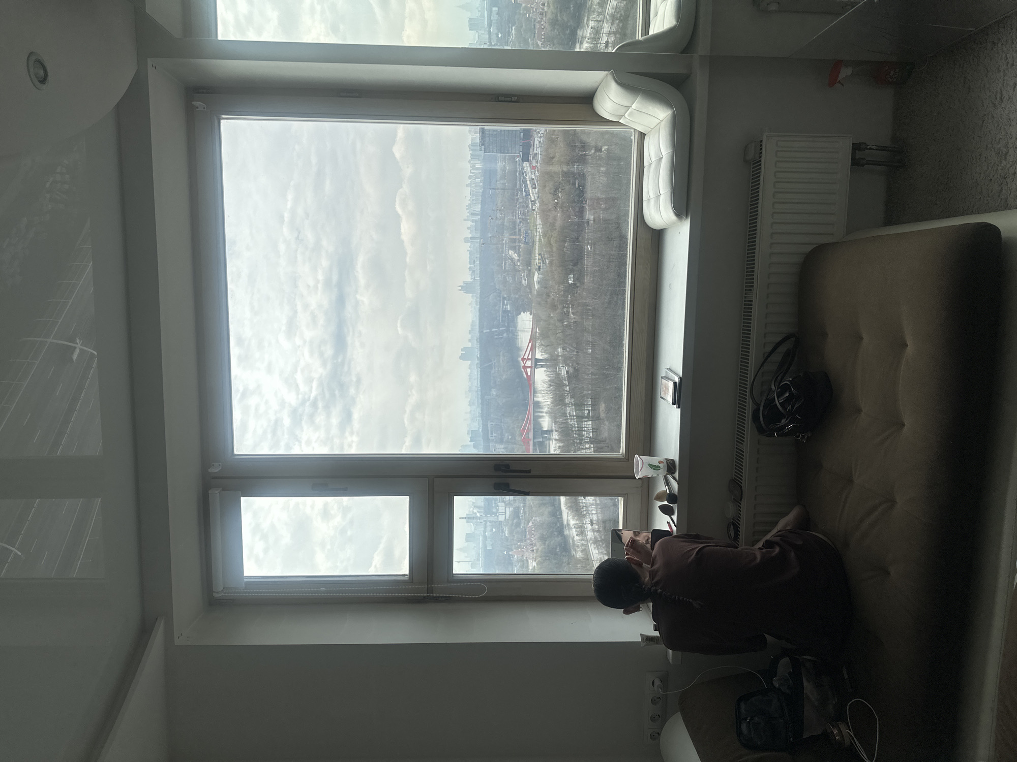 A person is sitting on a beige couch in front of a large window, looking at their phone, in an interior space. The window provides a view of a city skyline, partially obscured by clouds. The city appears to be Khimki, Russia. The view features buildings, trees, and a red cable-stayed bridge. A radiator is visible between the couch and the window. Makeup brushes and products are on the window sill. Other items, including a black bag, are on the couch and floor.
