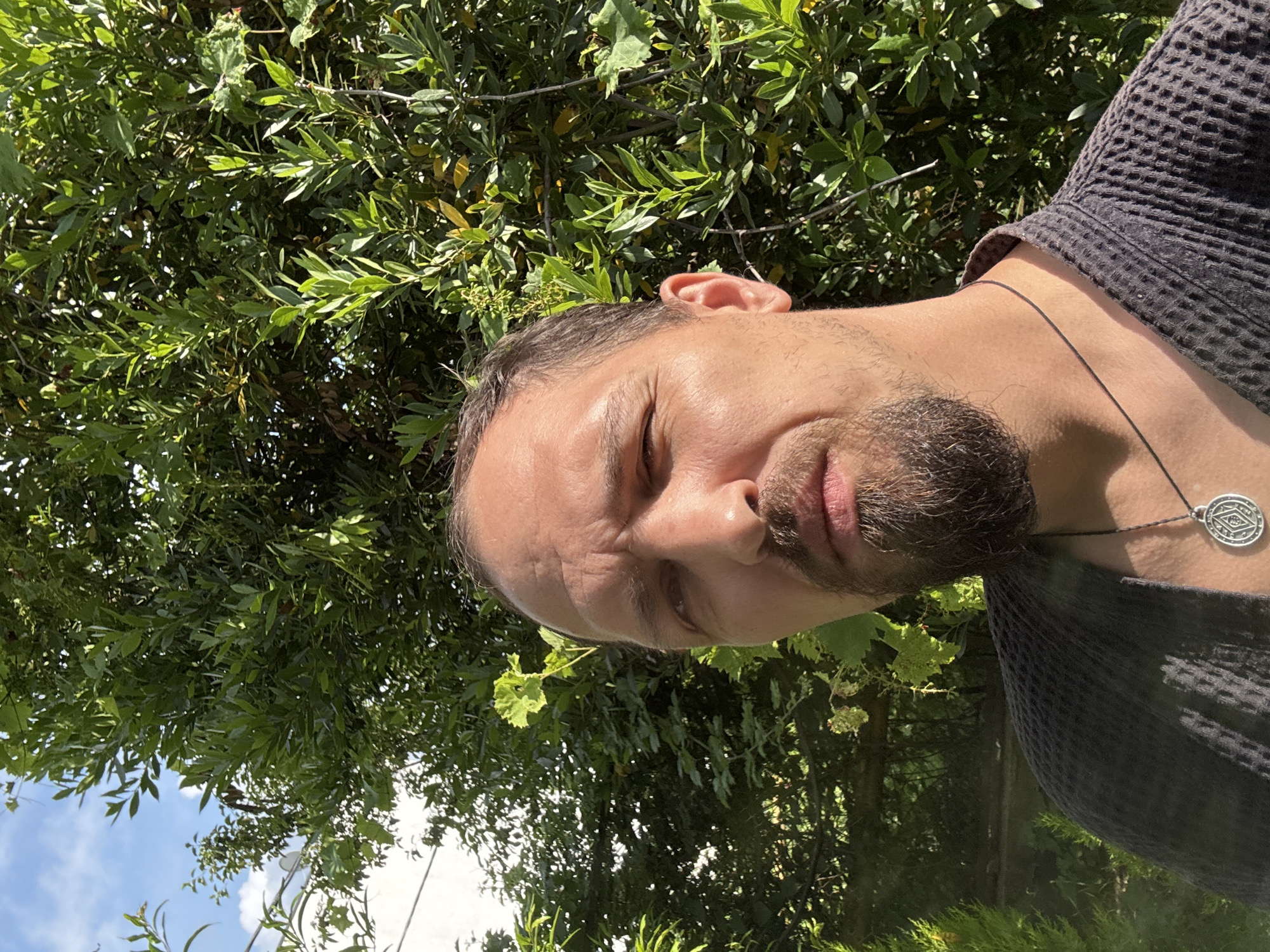 A man with a beard and a goatee, wearing a dark gray waffle-weave robe and a necklace with a circular pendant, is captured in a selfie outdoors. He stands in front of a lush green tree and a blue sky with some clouds. To the left of the frame, a white building and some wires are visible. The lighting suggests it is daytime. The scene appears to be in a residential or garden area, consistent with the city of Sochi, Russia, which is known for its abundant greenery and pleasant climate.