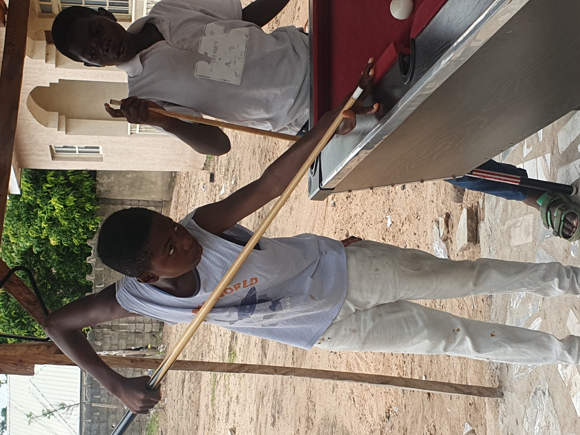 Two young males are engaged in a game of billiards outdoors in Lokoja, Nigeria. The foreground features a young male, wearing a light blue tank top with a graphic print and light-colored pants, in the process of striking a billiard ball with a cue. His stance suggests he is mid-shot, with his body angled towards the table. Beside him, to his right, stands another young male in a white polo shirt, observing the game. The billiard table is red and appears to be situated on a dirt surface. In the background, a building with an arched entrance and windows is visible, along with some vegetation and a fence. The overall environment suggests a casual, informal setting, possibly a backyard or a local recreational area.