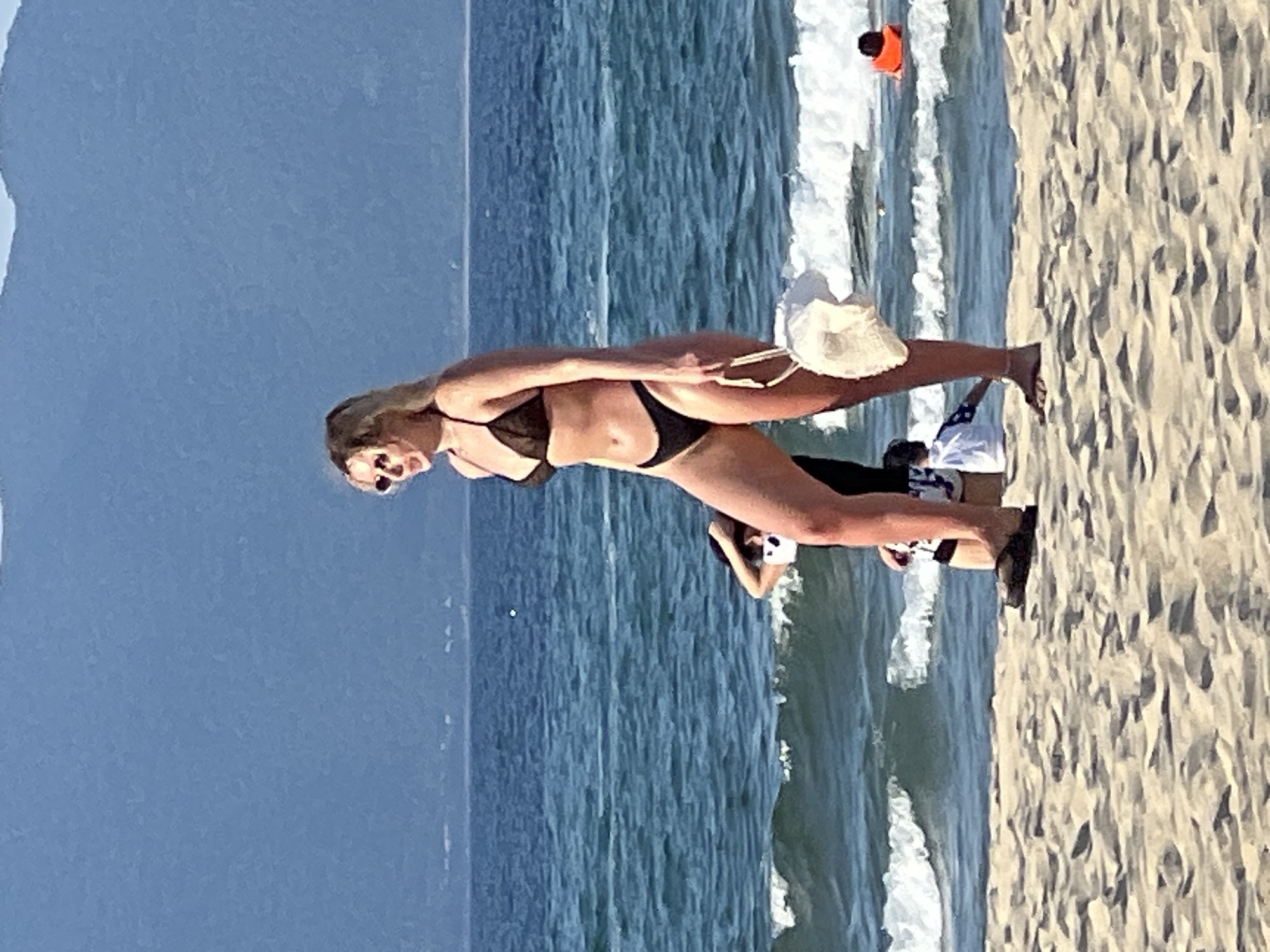 A woman with blonde hair, wearing a black bikini and sunglasses, stands on a sandy beach. She is holding a white woven bag. In the background, the ocean has waves, and distant mountains are visible. Other people are present on the beach and in the water, including a person wearing an orange life vest. The scene appears to be located on My Khe Beach in Đà Nẵng, Vietnam, known for its scenic coastline.