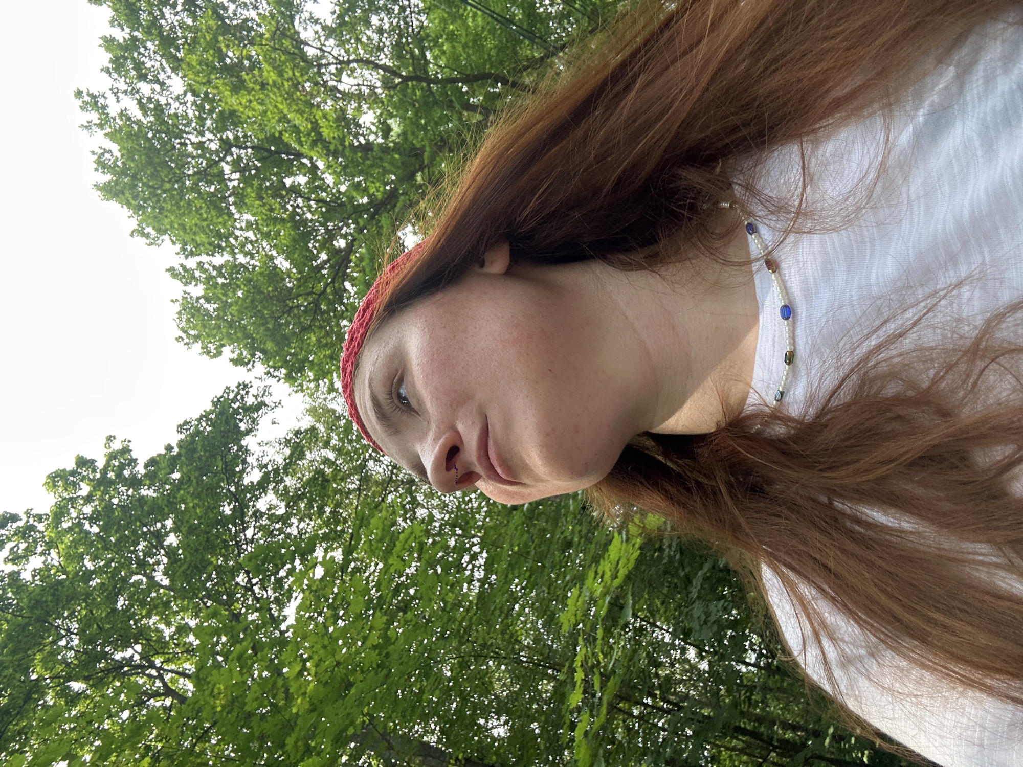 The image shows a close-up, low-angle shot of a person wearing a red headband and a white t-shirt. The person has long, reddish-brown hair and is looking upwards and to the left. A beaded necklace is visible around their neck. The background consists of green trees and a bright sky, suggesting an outdoor setting. There is no discernible connection to the city of Nizhny Novgorod, Russia.