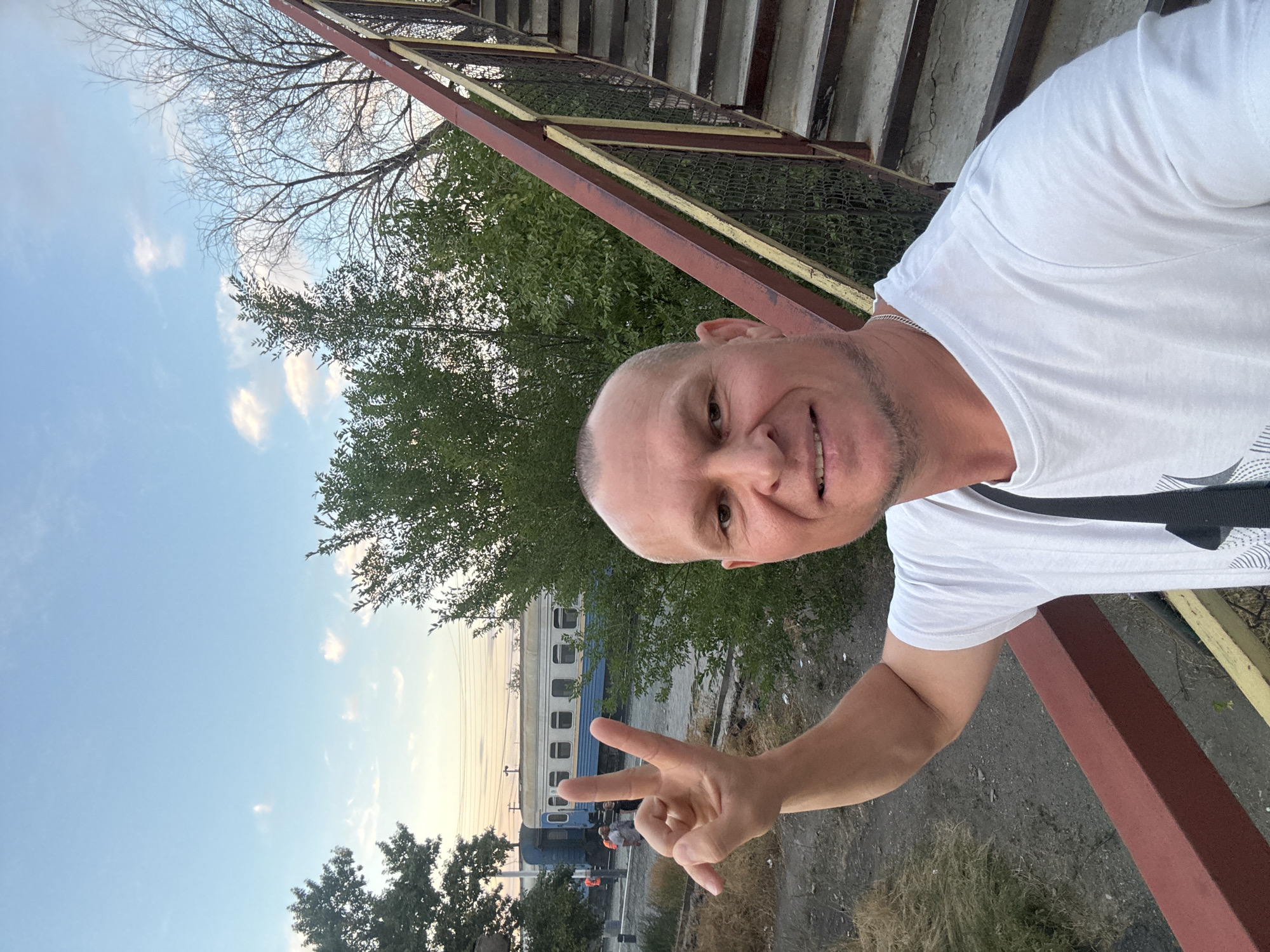A man with a shaved head and a white t-shirt is taking a selfie on an elevated walkway. He is smiling and holding up two fingers in a peace sign. In the background, a blue and white train is stopped on a track, and there are trees and a cloudy sky. The walkway appears to be made of metal and concrete. The man is wearing a silver chain around his neck and a dark bag strap across his chest. The overall scene suggests an outdoor, urban environment, possibly near a train station or industrial area.
