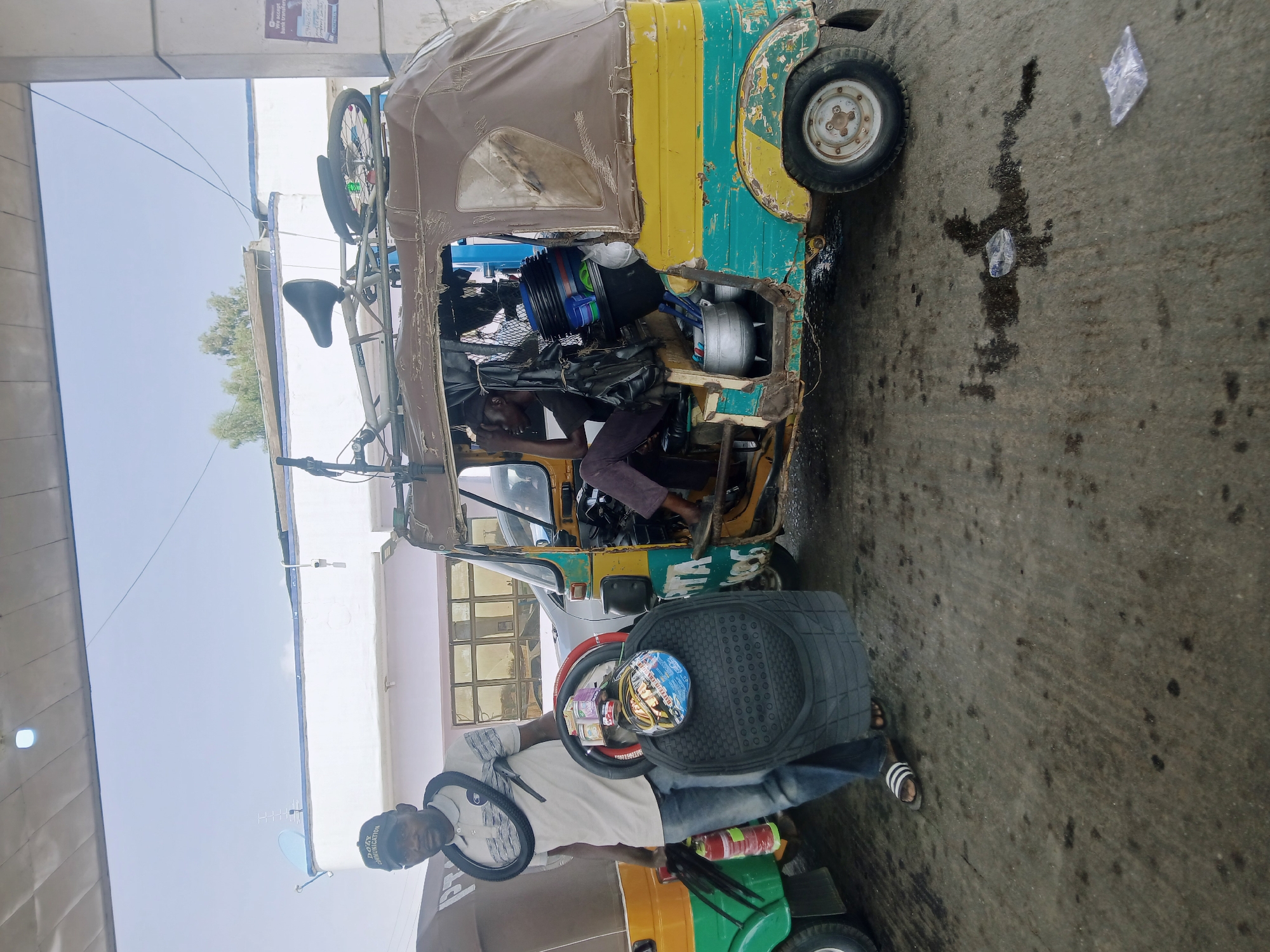 The image shows a scene in Jos, Nigeria. A brightly colored auto-rickshaw, filled with various objects including buckets, containers, and other unidentified items, is parked on a wet concrete surface. A bicycle is strapped to the top of the rickshaw. A man is seated inside the rickshaw, appearing to work on it. Another man stands beside a second rickshaw, holding a steering wheel. He is wearing a light-colored shirt, jeans, and sandals. A helmet is placed on the steering wheel in front of him. A green and yellow rickshaw is partially visible beneath the man holding the steering wheel. The surrounding environment includes building structures and a partially visible sky.