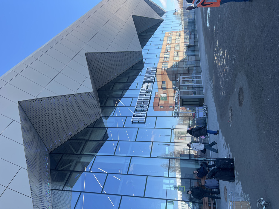 The image depicts the exterior of a modern airport terminal building under a clear blue sky in Murmashi, Russia. The building features an angular design with large reflective glass panels on the lower facade and light-gray metallic panels, some perforated, on the upper structure. A prominent sign on the glass reads "НА СЕВЕРЕ ЖИТЬ" in large white capital letters, with "ВХОД ENTRANCE" and an arrow below it, indicating the main entry.
In the foreground, a dark gray paved area shows two circular manhole covers. Several individuals are present:
* On the far left, a metal luggage cart is visible.
* Near the left side of the building, a wooden bench holds two seated individuals; one wears a dark jacket and beanie, the other dark clothing. Two dark wheeled suitcases rest on the pavement in front of them.
* A third individual, wearing a dark jacket and beanie, stands near the bench.
* Two females walk towards the right; one in a light jacket carries a dark shoulder bag, the other in a dark jacket has a pink item draped over her shoulder.
* Multiple silver luggage carts are parked near the glass entrance doors.
* On the far right, a person wearing an orange and black jacket is partially visible, cropped at the waist.
The glass facade reflects internal lighting, the blue sky, and an older, multi-story light-brown brick building located opposite the terminal.