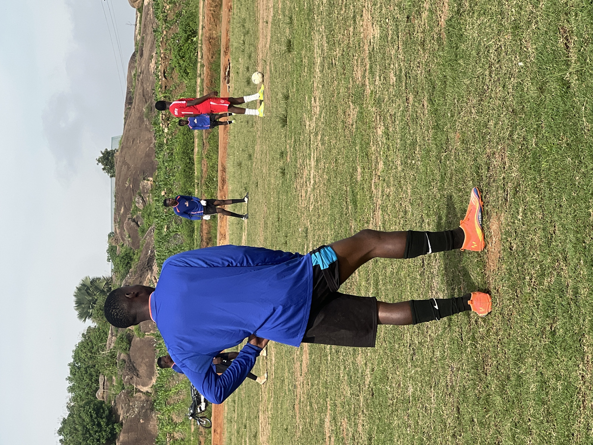 A group of young men are practicing soccer on a dirt and grass field, likely in the city of Jos, Nigeria. The foreground features a young man in a blue long-sleeved shirt and black shorts with blue accents, wearing orange soccer cleats and black socks. He stands with his hand on his hip, facing away from the camera. Behind him, other players are scattered across the field. One player in a red jersey and white shorts is near a soccer ball. Another player in a blue jersey and black shorts is further in the background. The environment consists of a somewhat uneven field with patches of grass and bare dirt. In the background, there are hills with rocky outcrops and vegetation, including trees and what appears to be a cornfield. A motorcycle is parked on the left side of the frame. The sky is overcast.