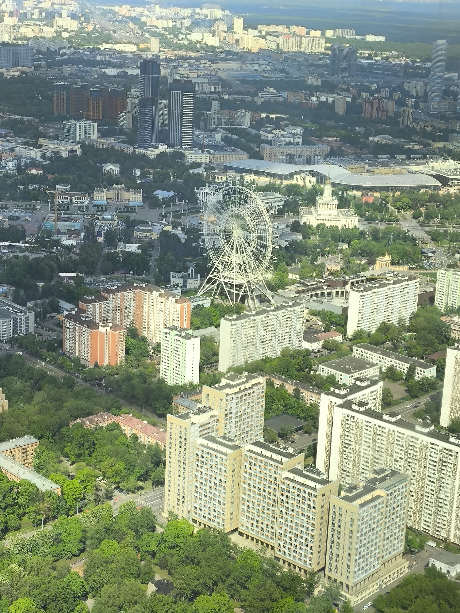 The image shows an aerial view of a city, with buildings, trees, and a large Ferris wheel in the midground. The cityscape extends into the distance, fading into a hazy skyline.