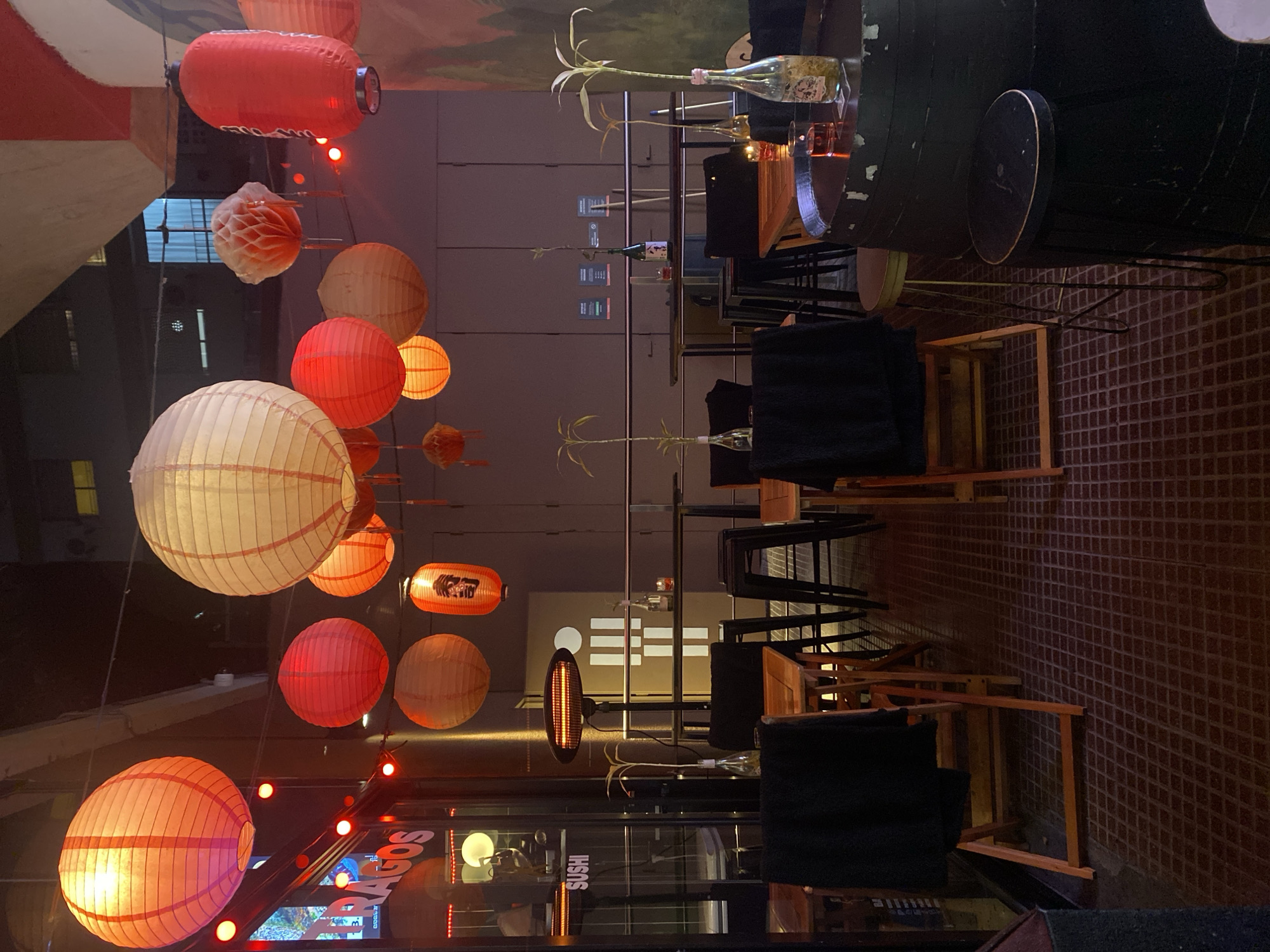The image depicts the interior of a restaurant, presumably in Buenos Aires, Argentina, based on the "BAGOS" sign. The restaurant is decorated with hanging paper lanterns in red and white hues. Wooden tables and chairs with black cushions are arranged in the dining area. Each table has a clear bottle with plant stems as a centerpiece. The restaurant has a modern interior with a visible storefront.
