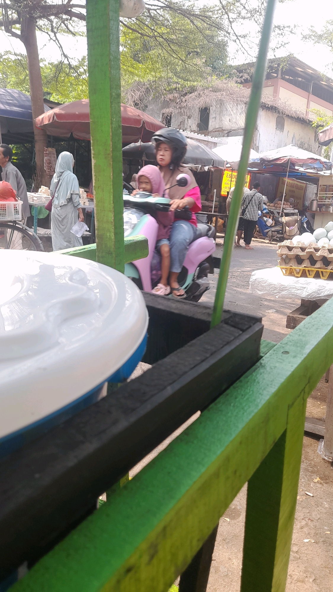 In Kepatihan, Indonesia, on a sunny day, a woman wearing a black helmet and a pink shirt rides a pastel-colored scooter. A young child, dressed in a pink hoodie, sits in front of her. They are passing by market stalls. One stall displays an assortment of eggs in cartons. In the foreground, a large white container is visible, possibly used for storing goods or drinks, with a blue base. The market appears to be outdoors, with trees and buildings in the background. The overall scene suggests a typical day in a bustling Indonesian marketplace.