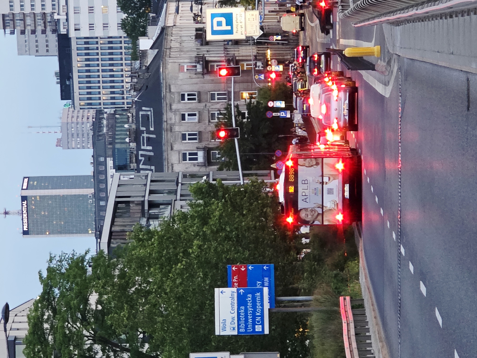 The image shows a street scene in Warsaw, Poland, at dusk. A bus with the advertisement "APLB" and license plate "WZ 46624" is stopped in traffic. Several other cars are also in traffic. Traffic lights are red. A parking sign is visible on the right side of the image. In the background, there are several buildings, including the Presidential Tower. Trees and foliage are visible on the left side of the image.