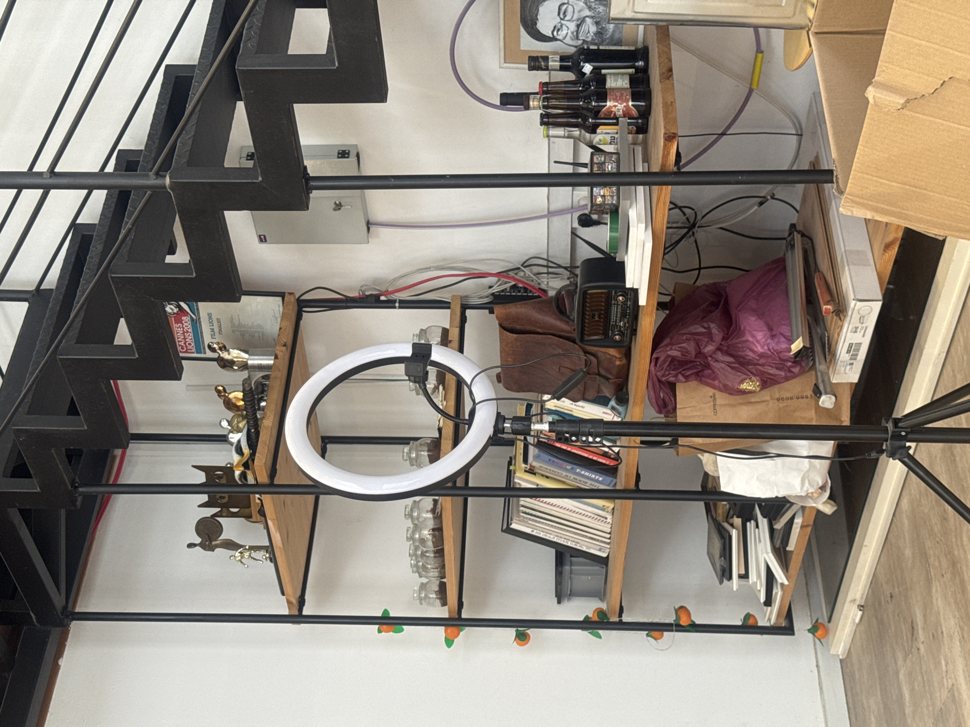 This indoor shot shows a cluttered space with several objects and architectural features. A black metal staircase runs diagonally across the left side of the image. To the right of the staircase, a wooden shelf unit is crammed with items including bottles, books, trophies, and a brown leather bag. A ring light is mounted on a stand in front of the shelves. A cardboard box partially obstructs the upper right of the frame. There is a portrait on the shelf, and some text elements are discernible, including "CANNES LIONS 2008 FILM LIONS FINALIST", "THE BOOK OF T-SHIRTS", "COFFEE", and "COFFEE...". The location appears to be an indoor space with evidence of storage and clutter, details such as the city of Tel-Aviv, Israel cannot be confirmed from the image.
