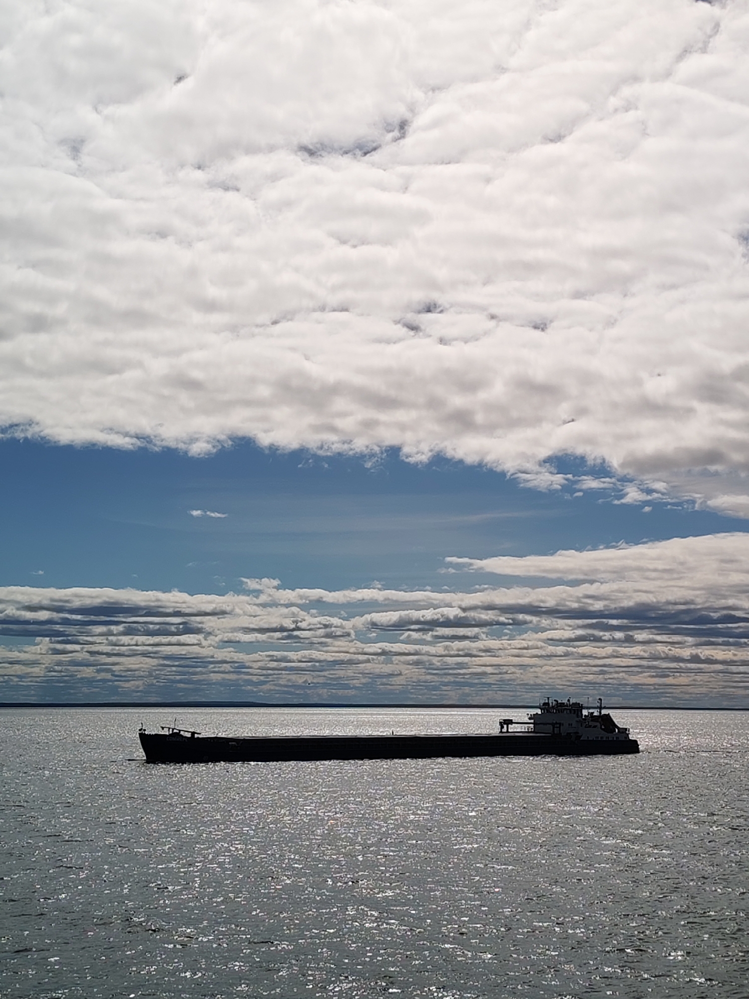 The image shows a long, dark barge on a body of water, likely Lake Beloye in Belozersk, Russia. The sky is predominantly covered with layered, cumulus clouds, with patches of blue visible. The water is shimmering with sunlight.