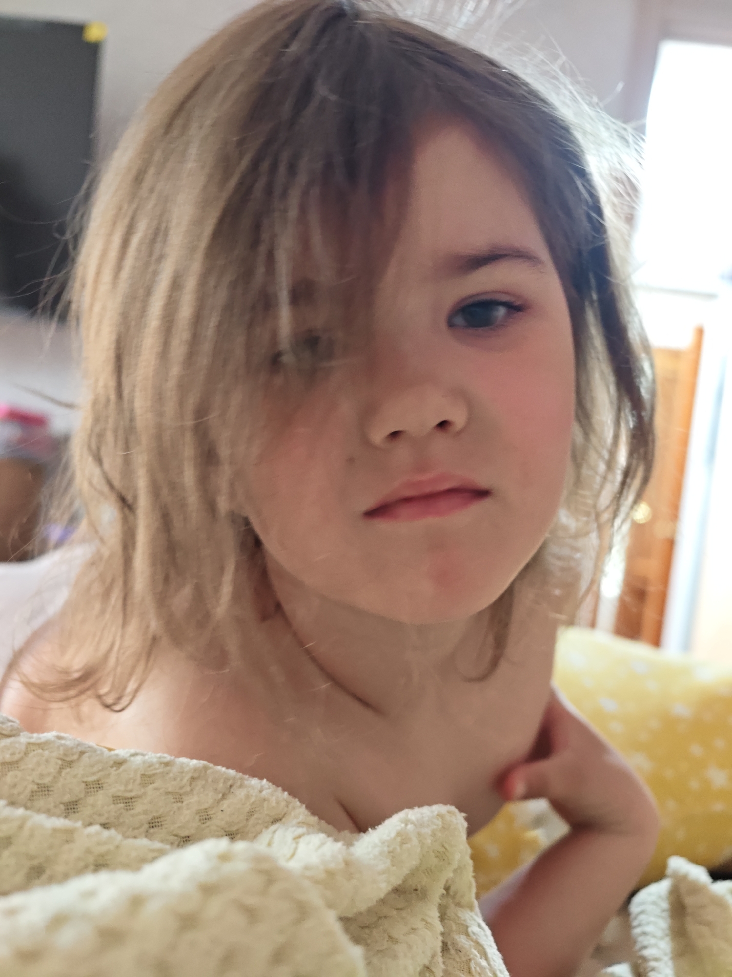 A close-up, slightly off-center shot captures a young girl with light brown, disheveled hair partially obscuring her face. She has fair skin, and a serious expression. The girl is wrapped in a soft, textured, cream-colored blanket. A yellow pillow with white star-shaped patterns is visible behind her right shoulder. The lighting suggests daylight. The location cannot be determined.