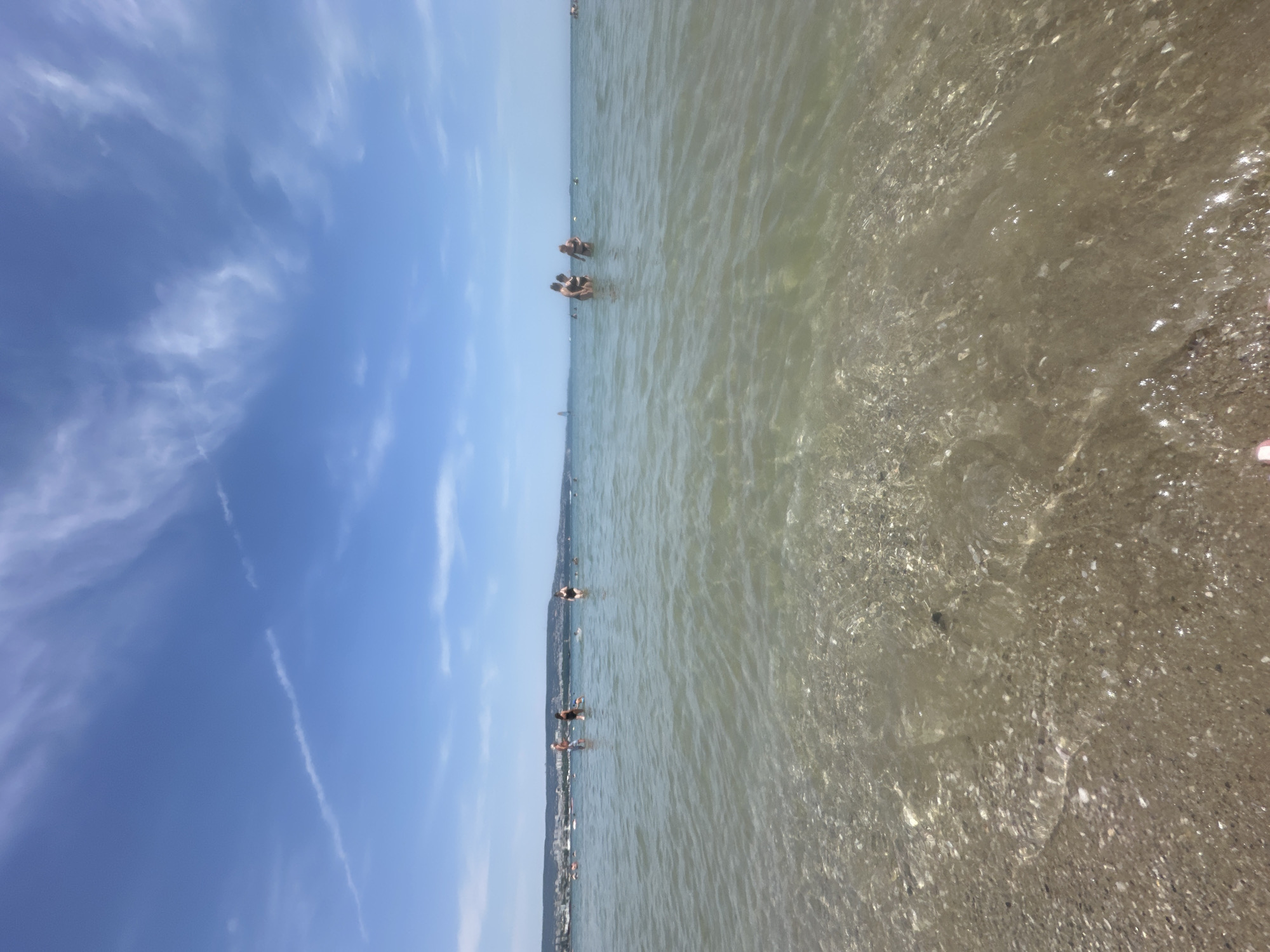 A wide-angle, low-perspective view of the Black Sea beach in Varna, Bulgaria, shows a sandy shoreline with clear, shallow water. Several people are wading and swimming in the water. In the mid-ground, a group of three individuals are standing in the water, and further to the left, a few more people are visible. The horizon displays a distant shoreline with buildings and greenery, indicating the city's presence. The sky is blue with scattered white clouds, and a thin contrail stretches across the upper portion of the image. The water is calm, with sunlight reflecting off its surface and illuminating the pebble and sand bottom.