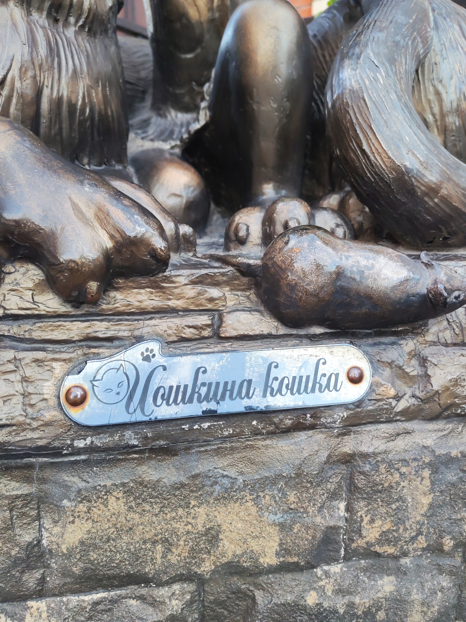 This media file shows a close-up of a bronze sculpture of cats and a mouse, located outdoors in Yoshkar-Ola, Russia. The sculpture is mounted on a stone facade, suggesting it is part of a larger public art installation or architectural feature.
The main subjects are the bronze figures of several cats and a mouse. The cats are depicted in various poses, with some appearing to be sitting or lying down. One cat's paw is prominent in the foreground. A mouse lies on its side in front of the cats.
A notable detail is a small, oval metal plaque attached to the stone below the sculpture. The plaque has a light blue background with black lettering and a simple outline of a cat's face and a paw print at the top. The visible text on the plaque reads "Кошкина кошка" which translates to "Koshkina koshka" or "The cat's cat." The plaque is secured with two small rivets.
The lighting suggests it is daytime, and the overall impression is of a well-maintained piece of public art. The texture of the stone and the bronze sculpture is detailed, showing the craftsmanship involved. The image does not provide specific clues about the time of day or weather conditions beyond what can be inferred from natural light. The overall context points to a city environment where art is integrated into public spaces.