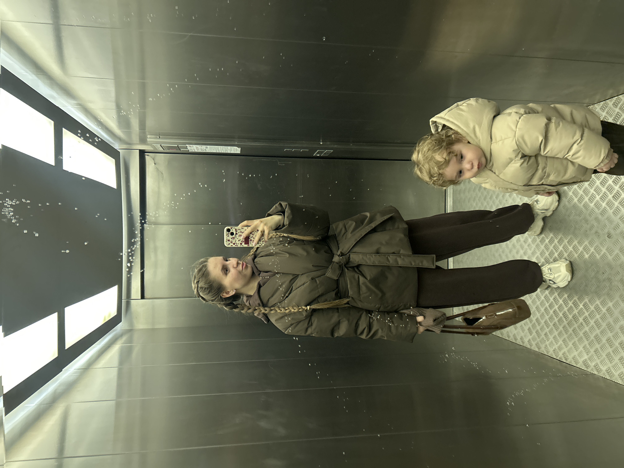 The image depicts a woman and a child taking a selfie in what appears to be an elevator with reflective metal walls. The woman, on the left side of the frame, is holding a smartphone and looking at it while extending her arm for the selfie. She is wearing a dark brown padded coat tied at the waist with a belt, and dark brown pants. Her hair is styled in a braid. She is also carrying a handbag.
To the right of the woman stands a young child wearing a beige padded jacket. The child is looking towards the camera. Both are wearing white sneakers.
The environment is a metal elevator with dim lighting, causing reflections and glare on the walls and floor. The floor is a metal grill texture. The location is not discernible from the image.
