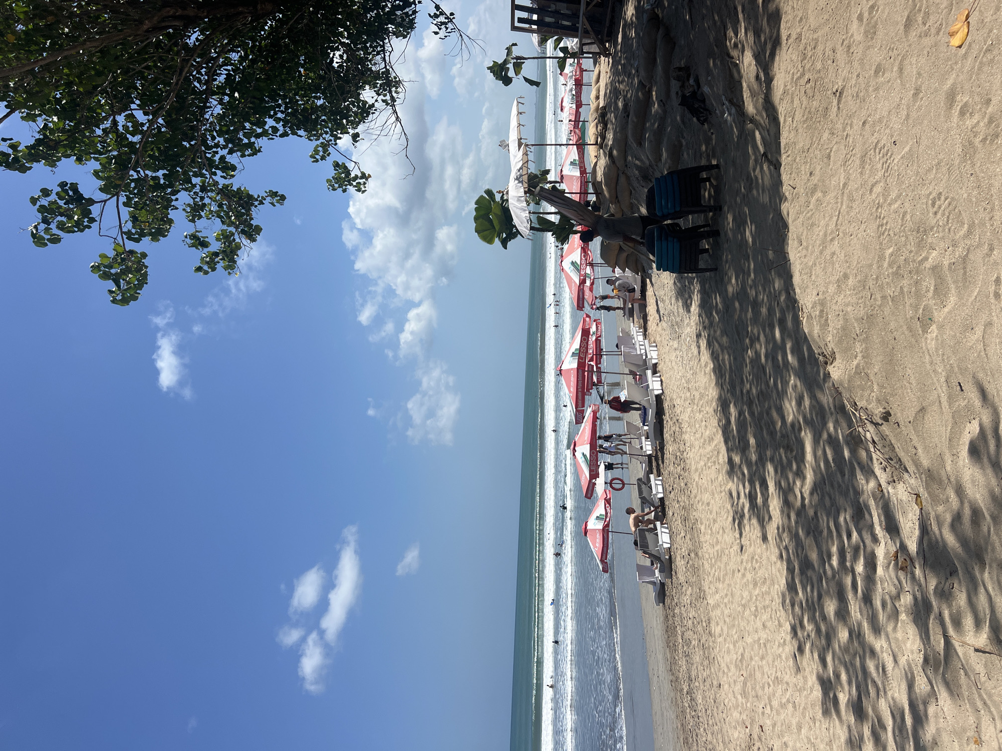 The image depicts a sunny day at Kuta Beach in Bali, Indonesia, with a wide expanse of sand leading to the ocean. Numerous beach chairs and red and white umbrellas are set up on the sand, many bearing the "LESSO" logo. Some beachgoers are visible relaxing on the chairs, while others are wading or swimming in the gentle waves. A few individuals are also seen standing near the umbrellas. In the background, surfers can be observed in the ocean. To the right of the frame, a large tree with green leaves overhangs the scene, casting dappled shadows on the sand. Several sandbags are piled up near some palm trees and a rustic wooden structure. The sky is a clear blue with a few white clouds. The overall atmosphere suggests a typical day at a popular tourist beach.
