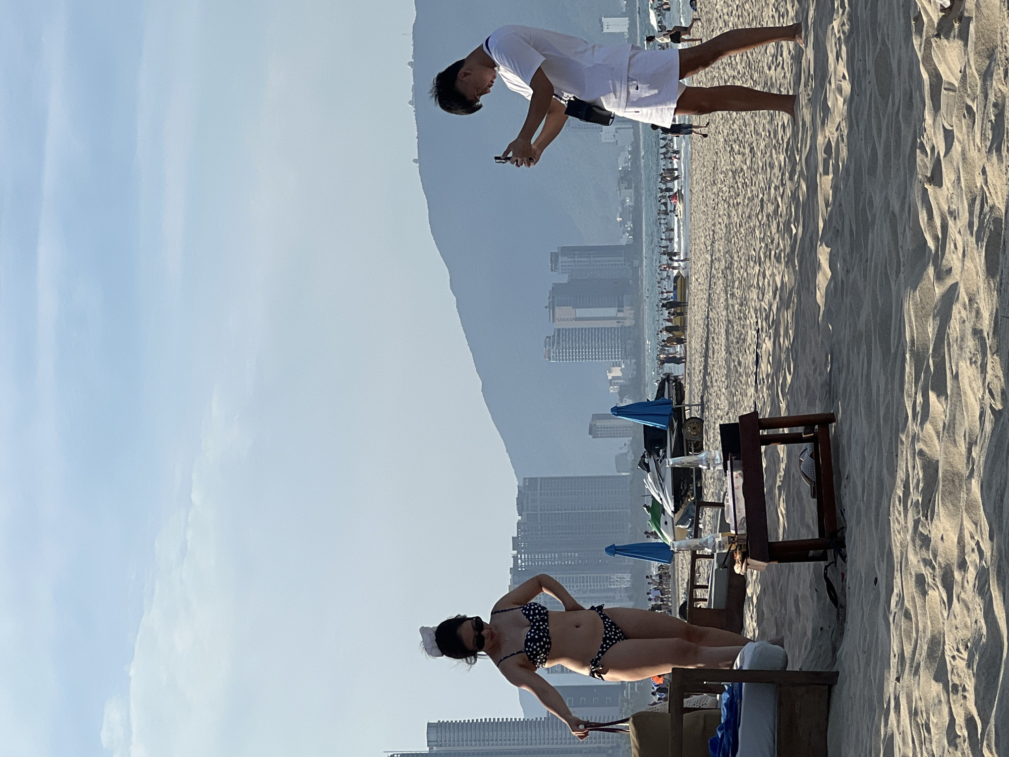 On a sandy beach in Đà Nẵng, Vietnam, a woman in a black and white polka dot bikini stands on the left, holding a purse. She is facing slightly away from the camera and has her left hand on her hip. In the middle ground, a man in a white t-shirt and white shorts is taking a picture with his phone. He is facing left and has a black bag slung over his shoulder. The background shows a crowded beach with many people in the water and on the shore. Several buildings and a mountain range can be seen in the distance. There are also some jet skis and umbrellas on the beach. The sky is clear with some scattered clouds.