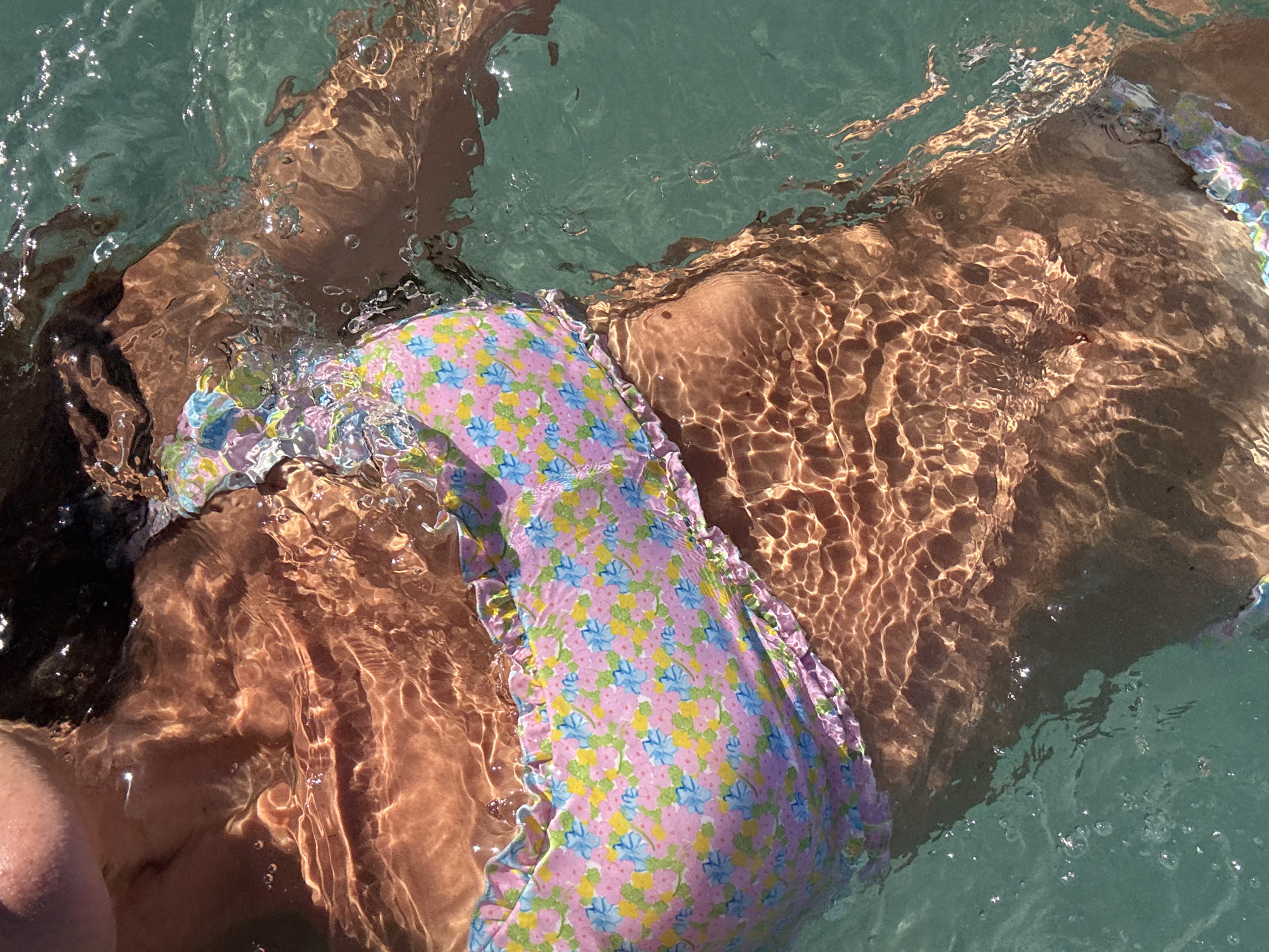 The image depicts a person in water. The subject appears to be submerged, with the body partially visible beneath the surface. The water is clear enough to allow some visibility of the person's torso and swimsuit, which is a light-colored floral print. The lighting creates a ripple effect on the body due to the water's surface. The location is labeled as the city of Unknown in Indonesia.