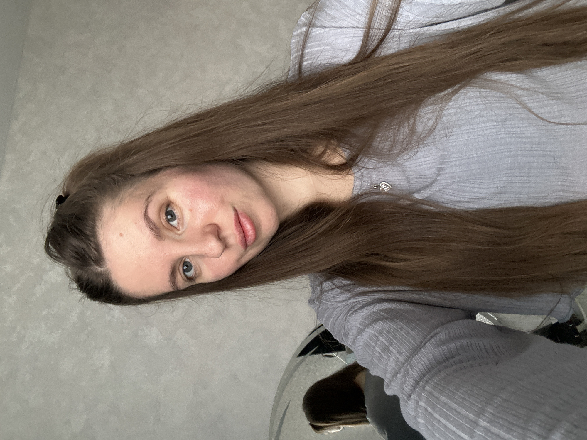 A woman with long brown hair and blue eyes poses for a selfie. She is wearing a light grey, long-sleeved shirt. A silver necklace is visible on her neck. The background appears to be a light grey wall. The location, Klin, Russia, cannot be determined from the image alone.