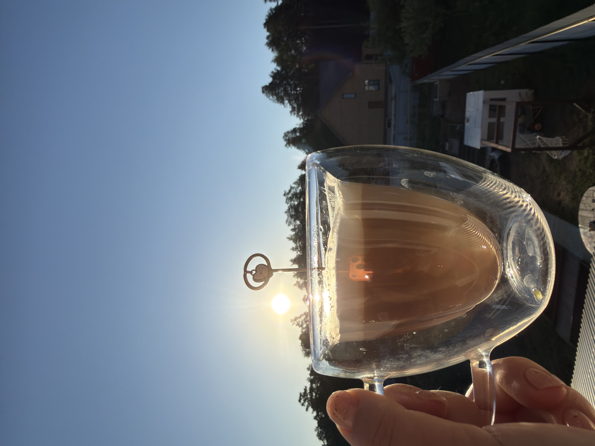 A medium shot, taken outdoors during daytime, shows a clear double-walled glass filled with a tan-colored liquid. An ornamental metal stick with a circular design at the top is placed inside the glass. A human hand is holding the glass. The sun shines through the glass, creating a halo effect around the circular design on the stick.
In the background, a view of trees, a house, and a small white building are visible. The sky is clear and light blue. The photo appears to have been taken in Chaikovsky, Russia.