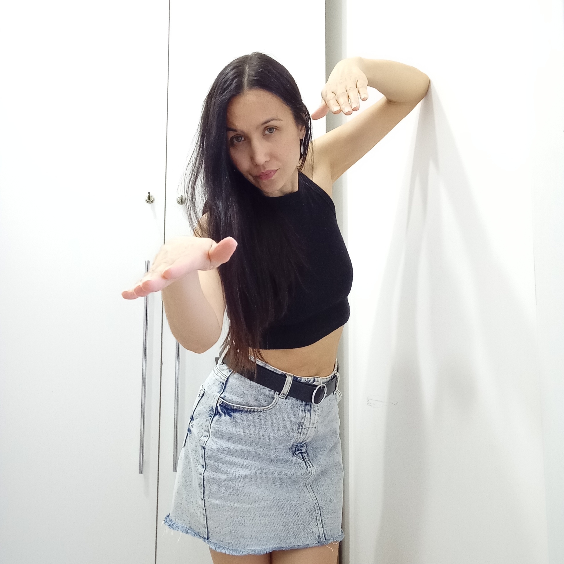 A woman with long, dark hair is posing indoors. She is wearing a black halter top and a light-wash denim mini skirt with a black belt. Her right arm is extended towards the viewer, palm open, while her left arm is bent with her hand near her head. She is leaning against a white wall and a white cabinet with silver handles is visible in the background. There is no specific evidence to indicate the location is Izhevsk, Russia.