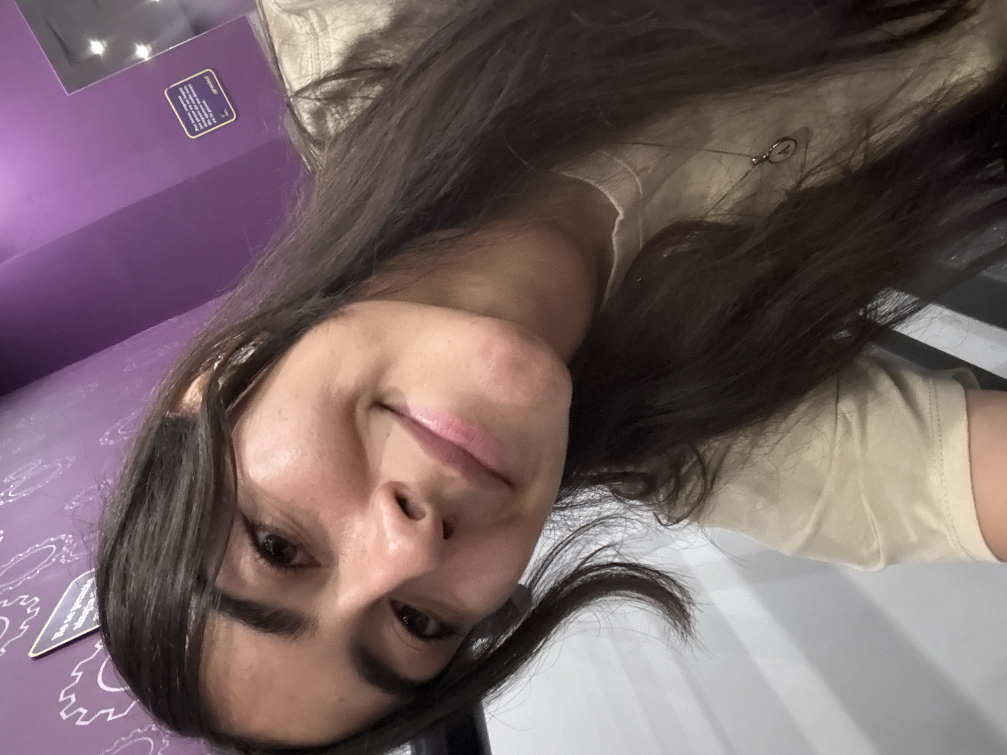 A young woman with long, dark brown hair and light-to-medium skin tones takes a selfie, looking down and slightly to her left with a faint, closed-mouth smile. She is wearing a light beige or tan-colored top. Her hair frames her face and flows over her shoulders.
The background on her left features a vibrant purple wall adorned with white, stylized patterns resembling gears or clockwork. On this wall, a small, dark rectangular informational plaque with white text is visible, though the text is largely unreadable due to blur and perspective. It appears to discuss something inspiring or historical. Above the plaque, a dark, reflective rectangular object, possibly a screen, reflects bright lights. A small "A" symbol is subtly visible within one of the white patterns on the purple wall near the bottom left. The background on her right consists of a plain white or very light-colored surface. A small, metallic loop or hook is visible near her collarbone, possibly part of an accessory or the top itself.
The setting appears to be an indoor public space, like a museum, exhibition, or themed attraction, given the decorated wall and informational sign. The woman's expression is pleasant and relaxed.