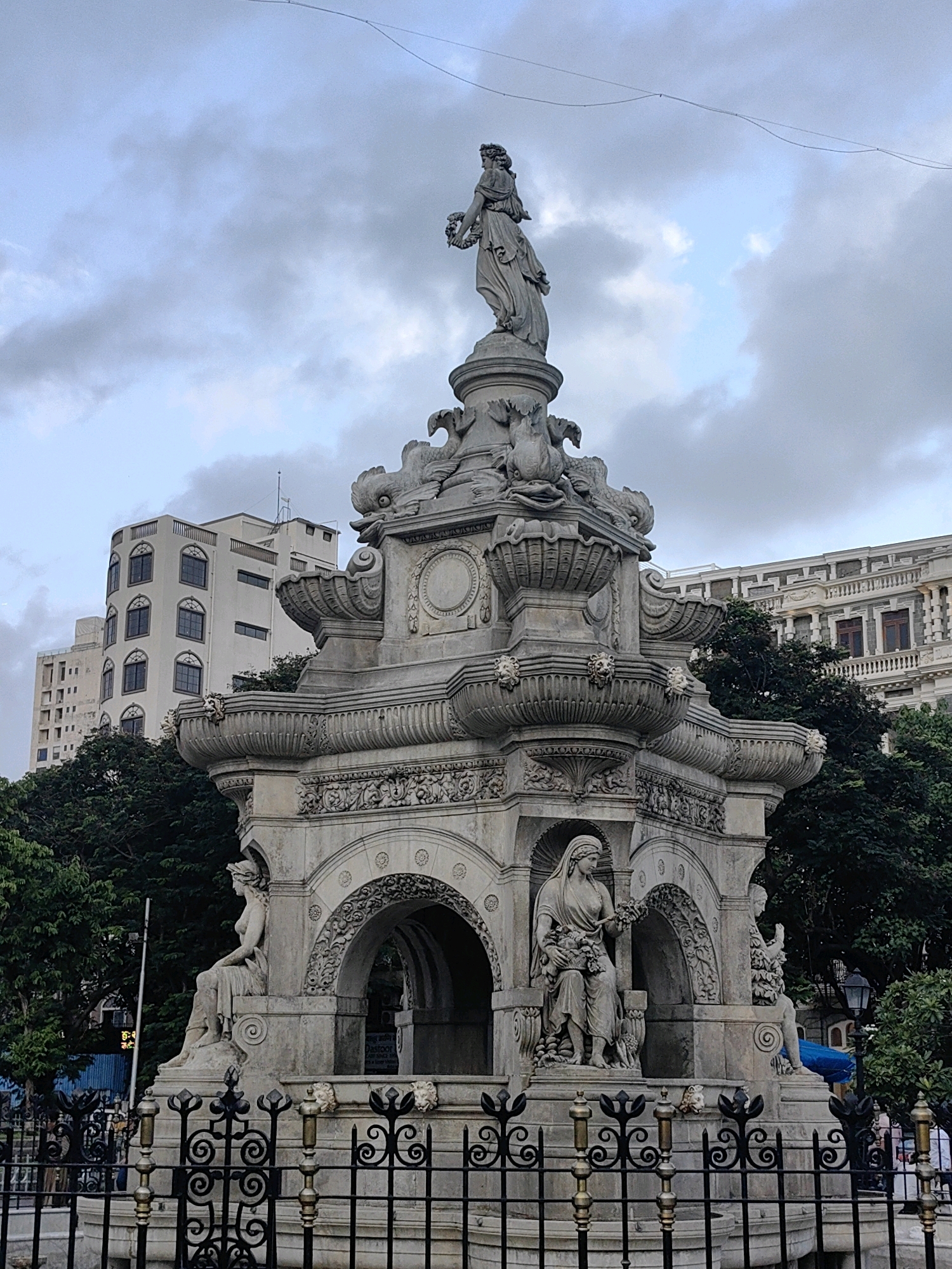 The image shows the Flora Fountain, a landmark in Mumbai, India, in a streetscape setting. The fountain is a tall, multi-tiered stone monument adorned with sculptures of female figures, mythical creatures, and decorative motifs. The main sculpture at the top depicts a woman holding a wreath. At the base of the fountain, there are seated statues of women, one holding a basket of flowers. The fountain is surrounded by an ornate black metal fence. In the background, there are several buildings of varying architectural styles, including a modern, multi-story building with arched windows and a more classical structure with ornate balconies and columns. Lush green trees are visible between the fountain and the buildings. The sky is overcast with scattered clouds. There are no visible people interacting with the fountain in the foreground. The scene appears to be an outdoor public space in an urban environment.