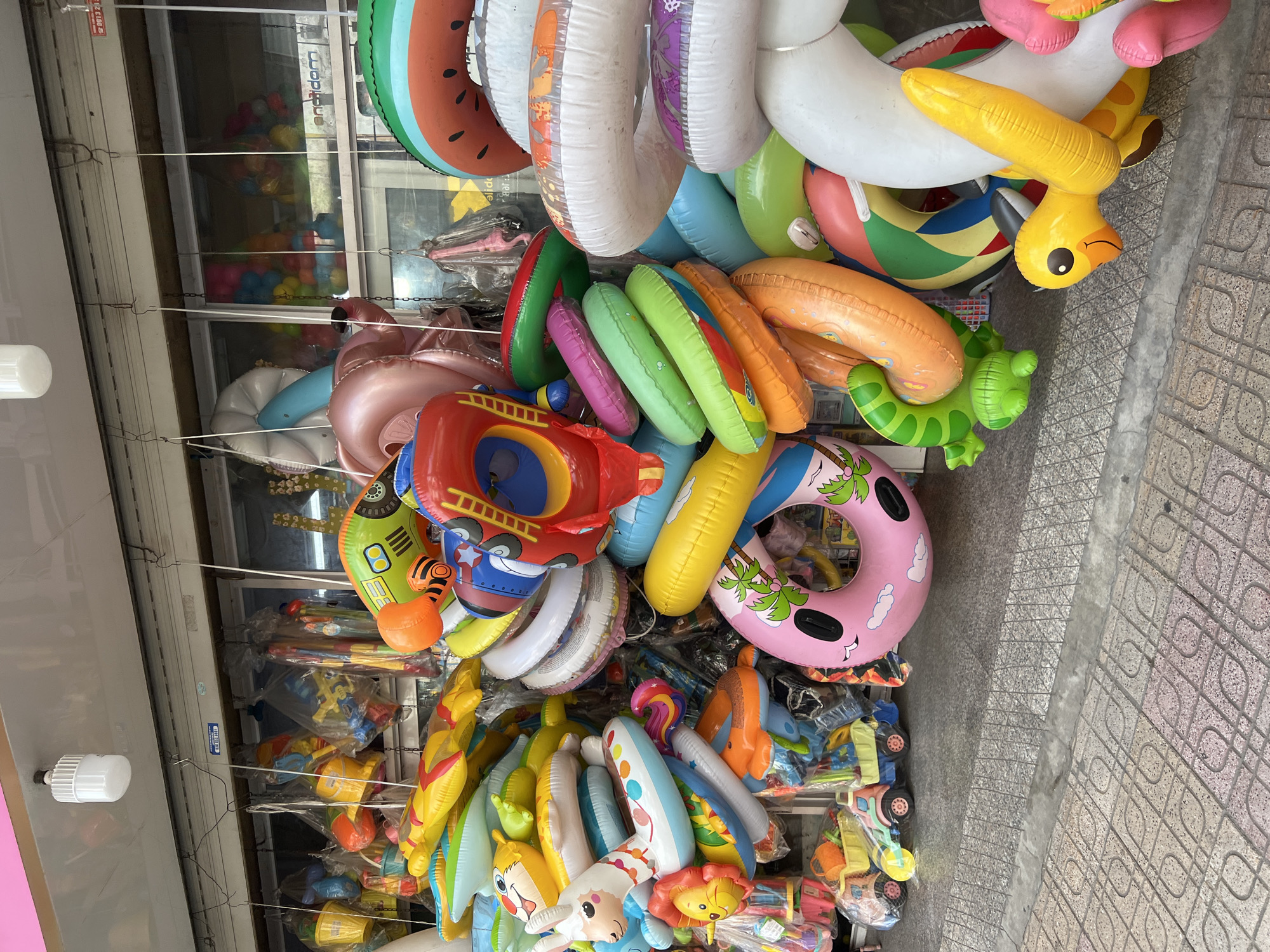 This is a full-shot, eye-level, outdoor, daylight photograph of a street-level stall in Nha Trang, Vietnam, displaying a large assortment of colorful inflatable toys, primarily swim rings and pool floats. The stall appears to be set up along a sidewalk with patterned stone paving. Behind the display, a metal security shutter is partially rolled up, revealing a shop interior with more merchandise visible through glass windows.
The foreground is dominated by a dense pile of inflatable objects. Among them are several large, circular swim rings, including one with a pink base featuring palm trees and clouds, another with a green and red striped design resembling a watermelon slice, and multiple white and translucent rings with patterned details. Scattered amongst these are various novelty inflatable toys: a yellow duck with prominent wings and beak, a green turtle with legs, and a red object resembling a rocket or fire truck with a ladder. There are also numerous other inflatable rings and shapes in shades of orange, yellow, green, pink, and blue, many with cartoonish designs of animals or characters. Some of these are stacked or leaning against each other.
Further back, more inflatable items are visible, including what appear to be inflatable boats or large floats, some with animal faces. Packages of toys, such as buckets and toy vehicles, are also displayed, particularly on the left side of the image. A white LED light fixture is visible hanging from the ceiling of the stall. The overall impression is one of a vibrant, playful commercial display, likely catering to tourists or local families looking for beach or pool accessories. No people are clearly visible within the frame of the photograph.