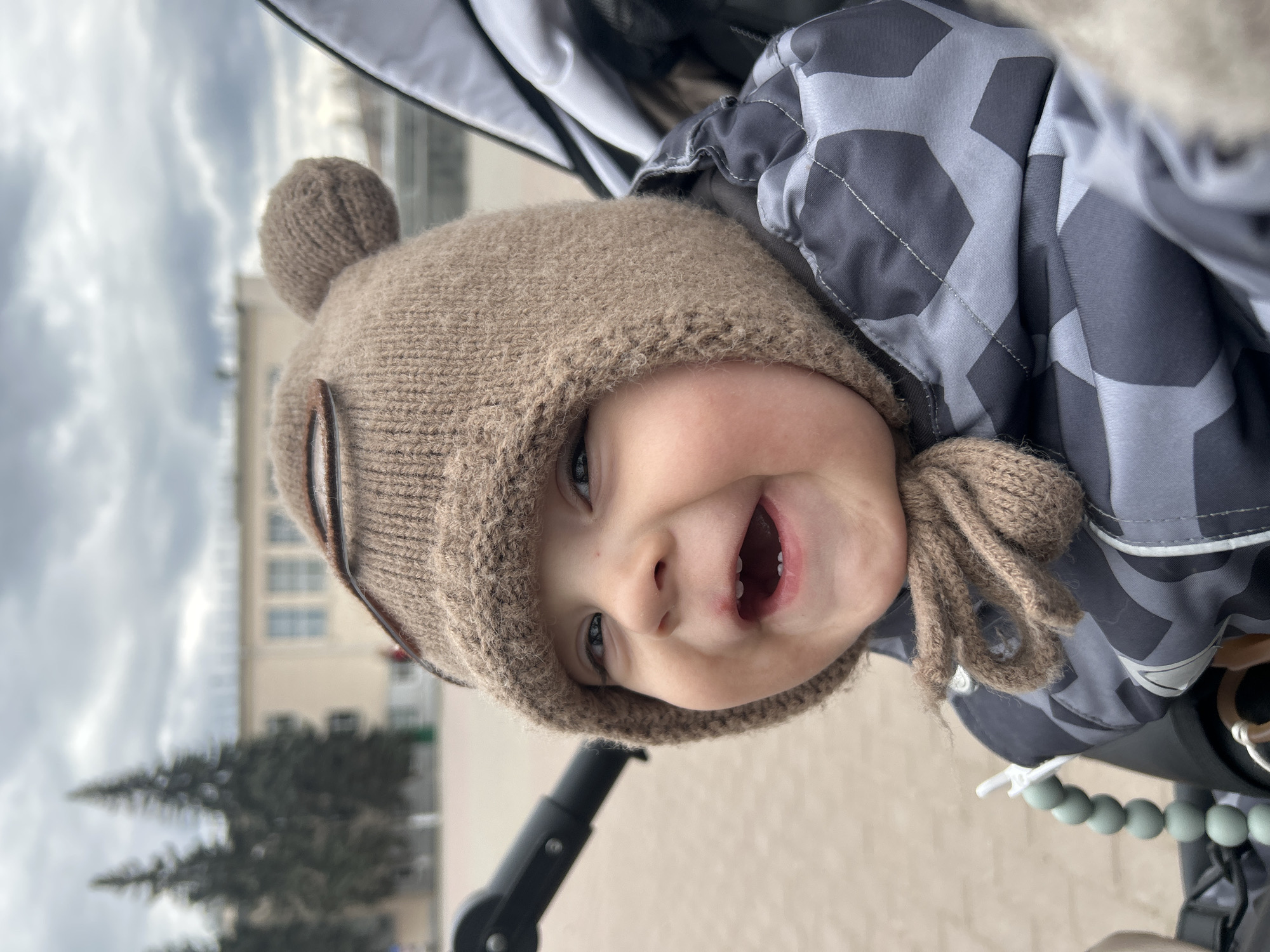 A happy baby, likely a toddler, is the central subject of this close-up image, smiling widely and laughing with an open mouth, showing some teeth. The child looks directly at the camera with bright eyes. They are dressed warmly in a light brown knitted hat with ear flaps and a small pom-pom on top, and a grey and dark grey patterned quilted jacket. Knitted braids or ties hang from the hat's ear flaps.
The child appears to be in a stroller or pram, with its dark fabric structure visible around them, and a black stroller handle or frame in the lower part of the image. A light green beaded teether or toy is clipped to the stroller's side.
The setting is outdoors in Chaikovsky, Russia, on what appears to be a cool, cloudy day, indicated by the overcast sky visible in the background and the child's warm clothing. Behind the child, a light-colored building with multiple windows and dark coniferous trees are visible. The ground in the foreground is light-colored and paved. There is no visible text in the media file.