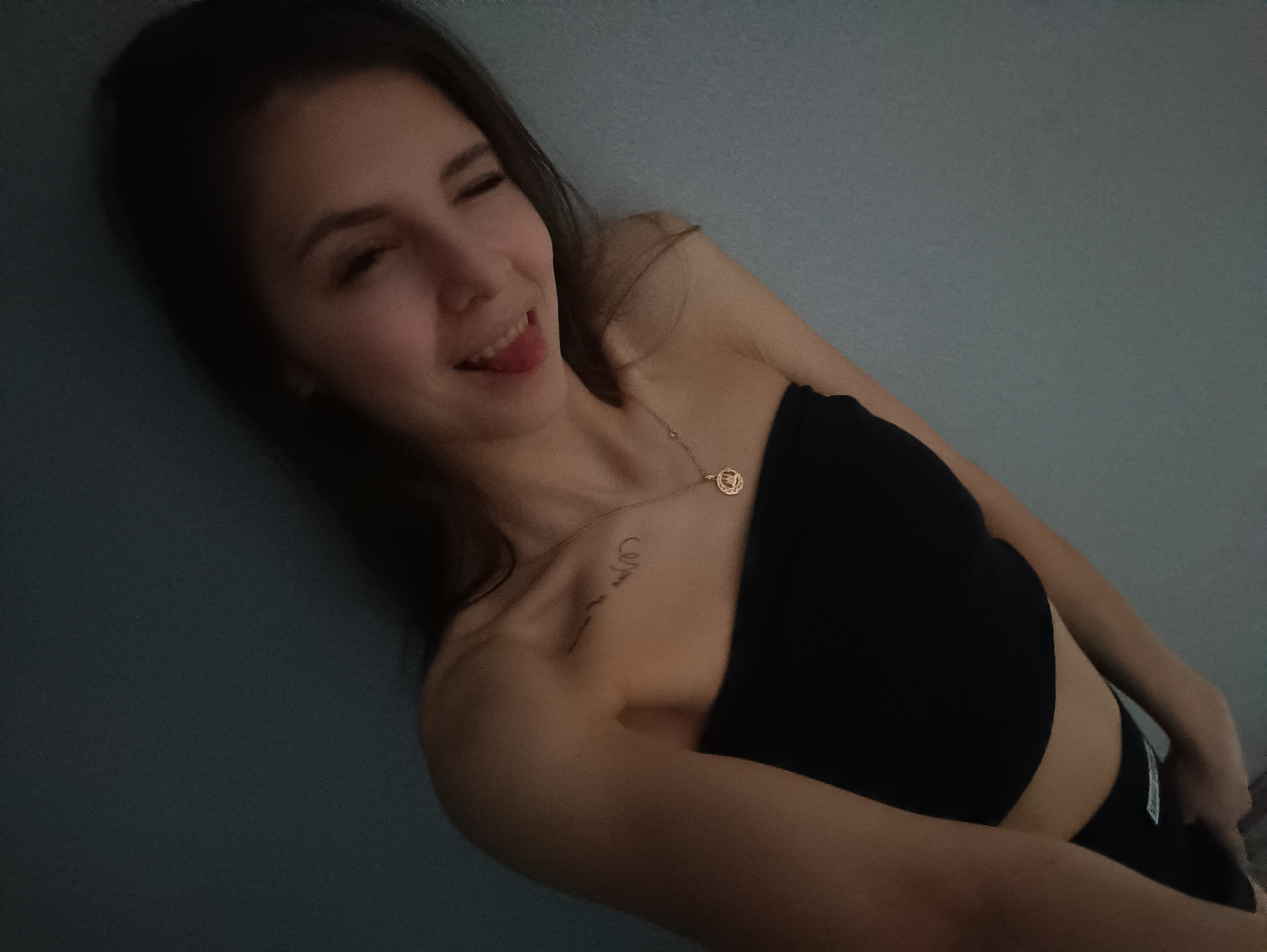 A woman lies against a bluish-gray wall. She is wearing a black tube top and black bottoms. She has a gold necklace with a circular pendant. A tattoo is visible on her upper chest/shoulder area. She is smiling with her tongue slightly visible. The photograph appears to have been taken indoors. There is no information to suggest the location is Chaikovsky, Russia.