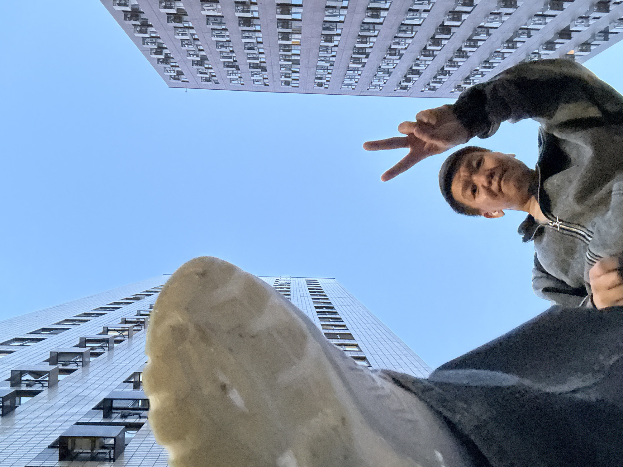 A low-angle shot captures a man making a peace sign towards the camera, with two high-rise buildings framing him against a clear blue sky. The building to his right has a regular grid of windows, while the building to his left is partially obscured by what appears to be a large, light-colored object, possibly a statue or part of a building. The man is wearing a gray and black jacket and has short dark hair. Based on the architectural style of the buildings, this scene may be in Moscow, Russia, which is known for its tall, modern apartment buildings.