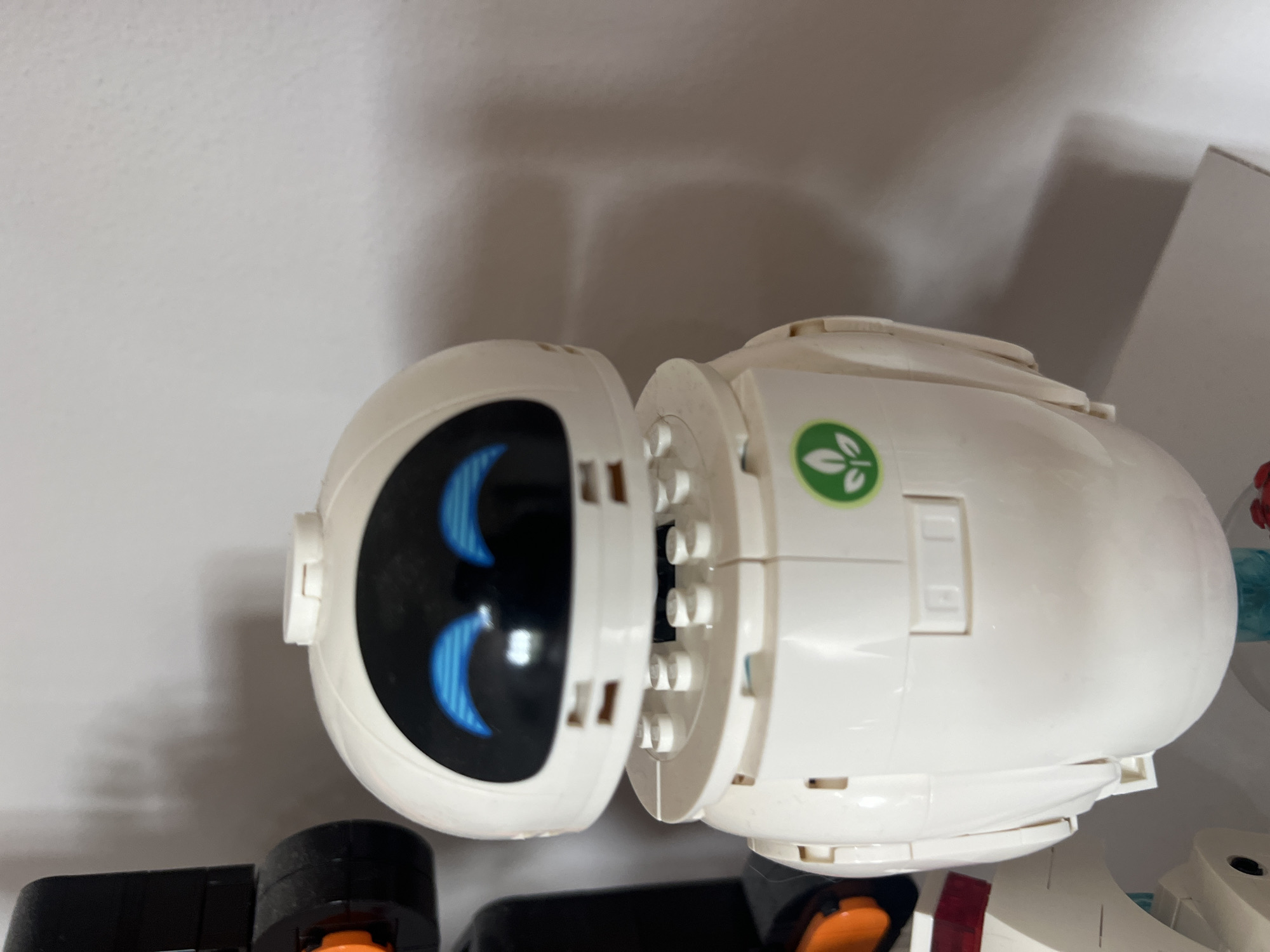 A close-up shot shows a white robot toy, likely constructed from LEGO bricks, positioned on a surface. The robot has a rounded head with blue, glowing eyes and a black faceplate. Its body is also white and appears to be assembled from modular pieces. A green circular emblem with a leaf design is visible on the robot's chest. To the left of the robot, a portion of a black toy structure with orange accents is visible. The background is a plain, light-colored wall with some subtle shadows, suggesting it is an indoor setting. There is no indication of the city of Onokivtsi, Ukraine, or any other specific location details within the image.