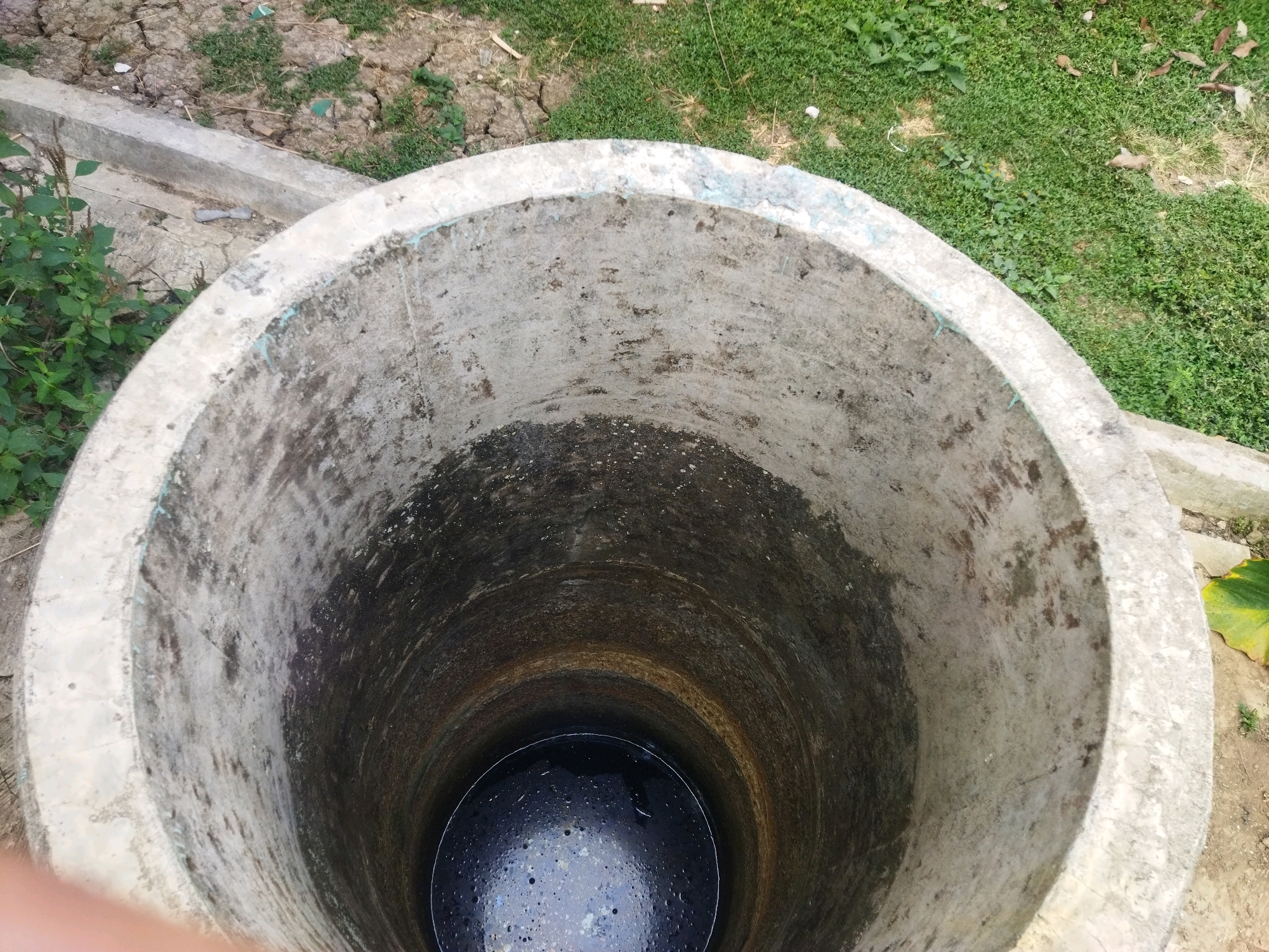 The image depicts a top-down view of a circular concrete well filled with dark water. The well's interior walls are stained and weathered. Surrounding the well is a dry, cracked earth area with patches of green grass and some scattered debris, including leaves and what appears to be a piece of light-colored plastic. A concrete edge or structure is visible to the left and right of the well opening. The overall impression is an outdoor, somewhat neglected environment, likely in a rural or semi-urban setting, consistent with the location of Kesugihan, Indonesia. There are no people or animals visible in the image.