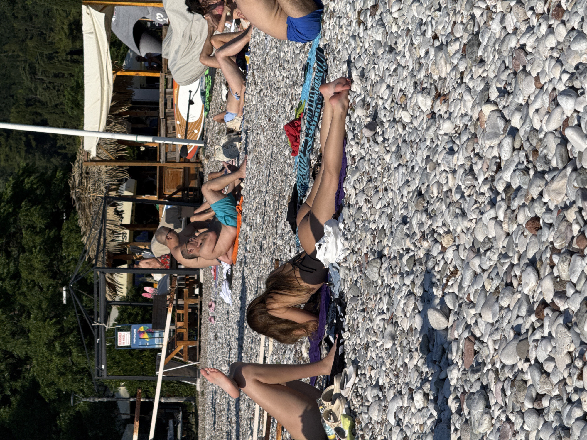 The image depicts a crowded pebble beach in Budva, Montenegro, with numerous people sunbathing and relaxing. In the foreground, a person's legs are visible, resting on a purple towel, with several pairs of sandals scattered nearby. Further back, a woman with long brown hair is lying on her stomach on a towel, facing away from the camera. Other individuals are visible in the middle ground and background, some lying on towels, others sitting or standing. Beach umbrellas and structures, including a thatched-roof kiosk and a white tent, are present. Several paddleboards are leaning against a structure in the background. The environment is characterized by a bright, sunny day, with shadows cast across the beach. The scene suggests a typical summer day at a popular tourist destination.
