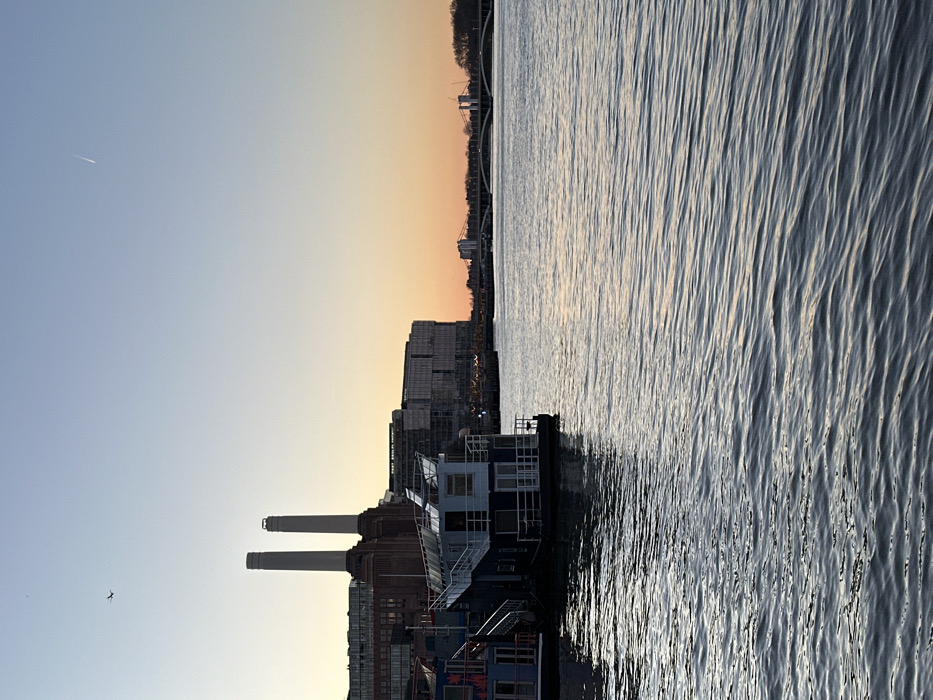 The image features Battersea Power Station along the River Thames. Two prominent chimneys stand out against the backdrop of a sunset sky, transitioning from blue to orange. A large body of water reflects light, creating a wavy pattern. Buildings and other structures line the riverbank, with a bridge visible in the distance. A few birds can be spotted in the sky.