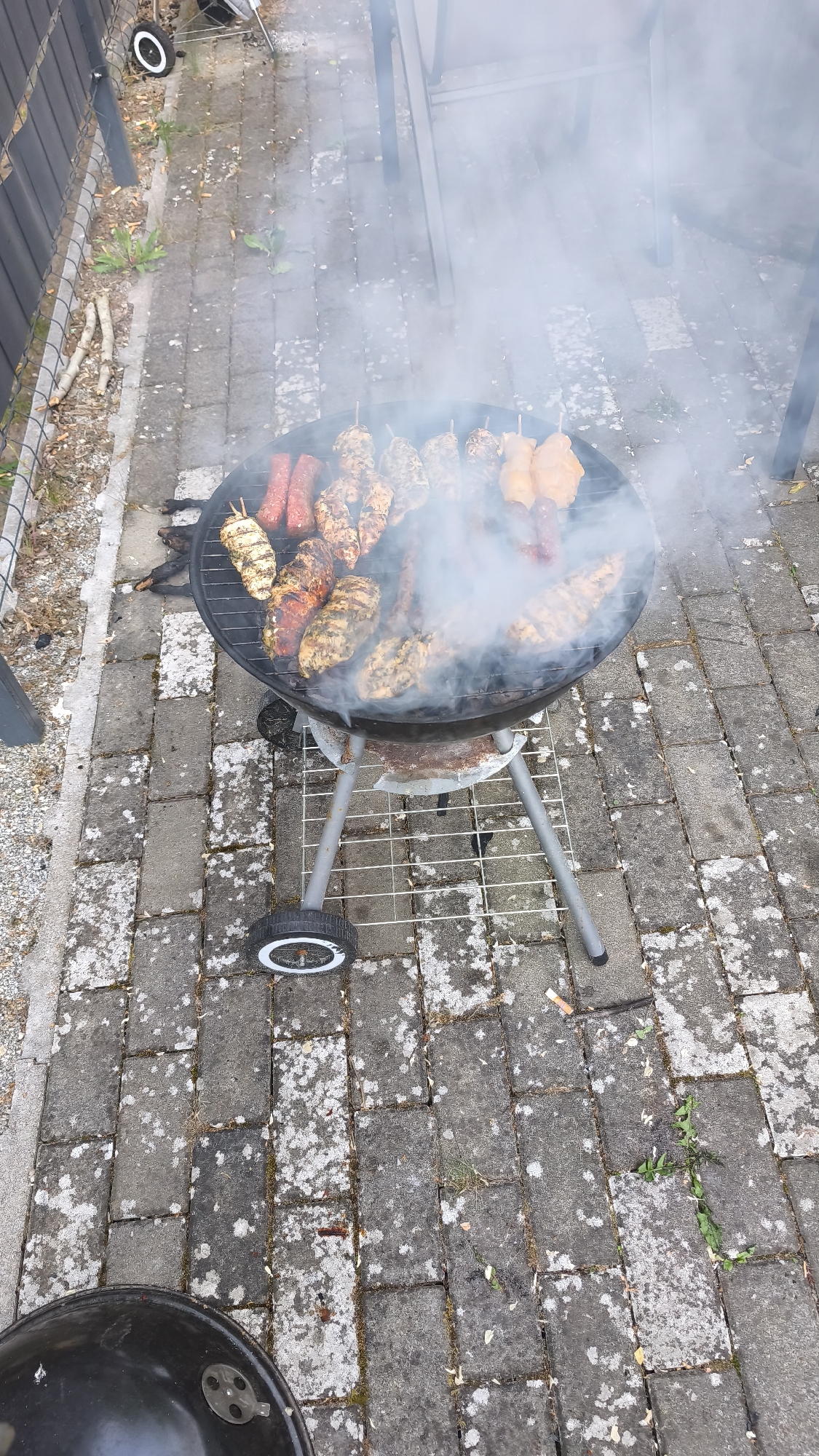 The image depicts an outdoor patio in Unknown, Unknown, featuring a black, circular, kettle-style charcoal grill actively cooking various meats. Dense white smoke rises from the grill, partially obscuring the background. On the grill grate, multiple pieces of poultry (chicken breasts/thighs, some on skewers) are visible, exhibiting grill marks and varying stages of doneness. Several red, cylindrical items, consistent with sausages or chorizo, are also cooking on the grate. The grill is supported by a three-legged metal stand, two of which have black wheels with white rims. A wire rack is positioned on the stand below the grill basin.
The ground is paved with rectangular grey pavers, exhibiting widespread white and green discoloration from lichen or moss, particularly within the mortar lines and on the paver surfaces. Scattered organic debris, including small leaves, twigs, and multiple discarded cigarette butts, is present on the pavement. To the left, a dark-colored fence, possibly metal, runs parallel to the grill, with a section of wire mesh visible near its base. Beyond the fence, an unpaved area with sparse vegetation and small branches is discernible. In the upper right portion of the image, the lower legs of light-colored outdoor furniture, likely a table and chairs, are partially visible through the smoke. The scene is illuminated by ambient daylight.