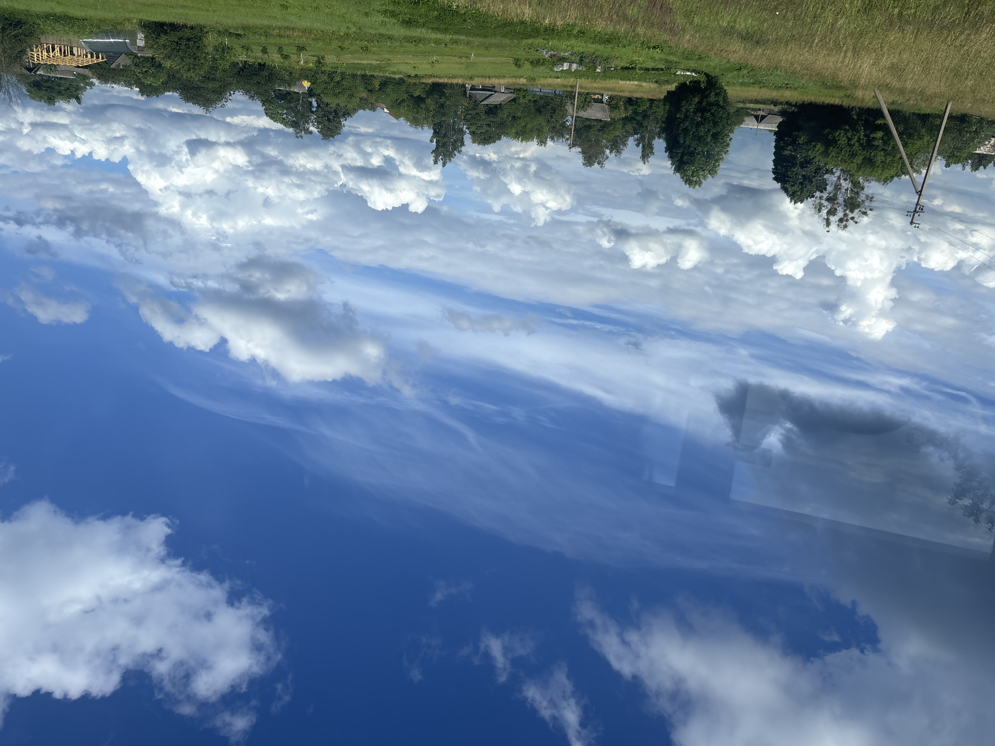 The image depicts a rural landscape under a dynamic sky with both wispy cirrus clouds and larger cumulus formations against a vibrant blue backdrop. The foreground is dominated by a green field with tall grasses, leading to a gentle slope. In the middle ground, a scattered collection of trees and vegetation borders a settlement. Several small, rustic buildings are visible, some appearing to be wooden houses with dark roofs, and one structure is in a state of construction with exposed wooden beams. A utility pole with wires stands prominently on the left side of the field, with its wooden support structure clearly visible. The overall impression is that of a tranquil, rural setting in the vicinity of Грозаўскі сельскі Савет, Belarus, likely during daylight hours with pleasant weather.