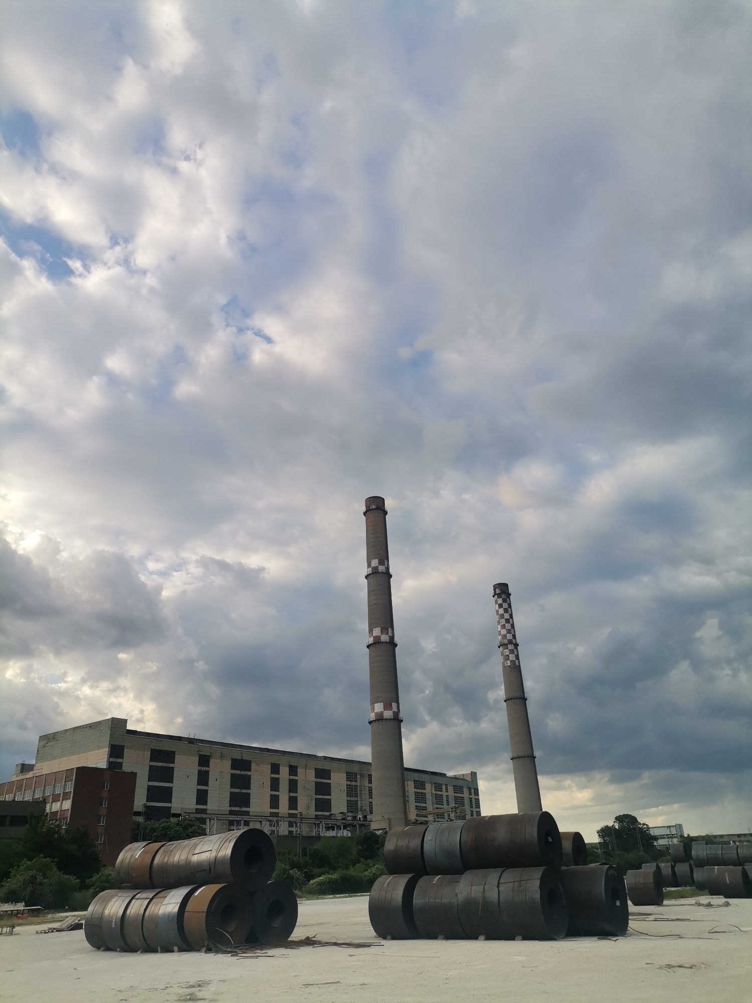 The image shows an industrial scene under a cloudy sky, likely taken in or near Ezerovo, Bulgaria, given the presence of industrial infrastructure typical of the region. Two tall, concrete smokestacks rise in the center of the frame, one with alternating red and white bands near the top and the other with checkered pattern markings. In the background, a large, multi-story industrial building with multiple windows. In the foreground, there are stacks of cylindrical metal coils stored on a flat, light-colored surface, likely concrete or packed earth. The coils appear to be rusty or tarnished. Sparse vegetation is present in the space between the stacks and the building. The scene conveys a sense of industrial activity and infrastructure.