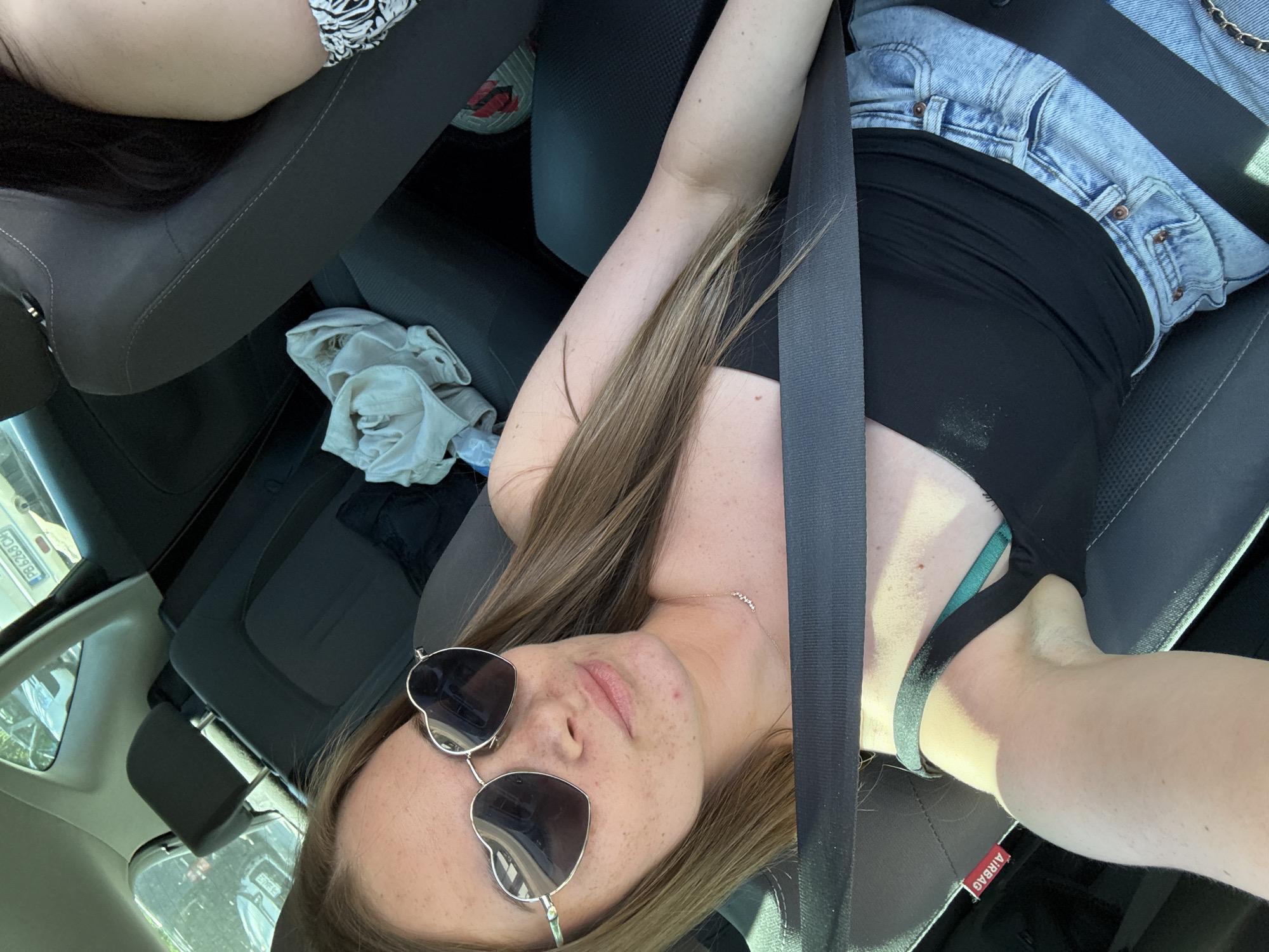 A young woman wearing heart-shaped sunglasses sits in the driver's seat of a car, secured by a seatbelt. She is wearing a black top and denim shorts. The car's interior is visible, including the dashboard and passenger seat. The car registration plate is visible from the windows in Plovdiv, Bulgaria.
