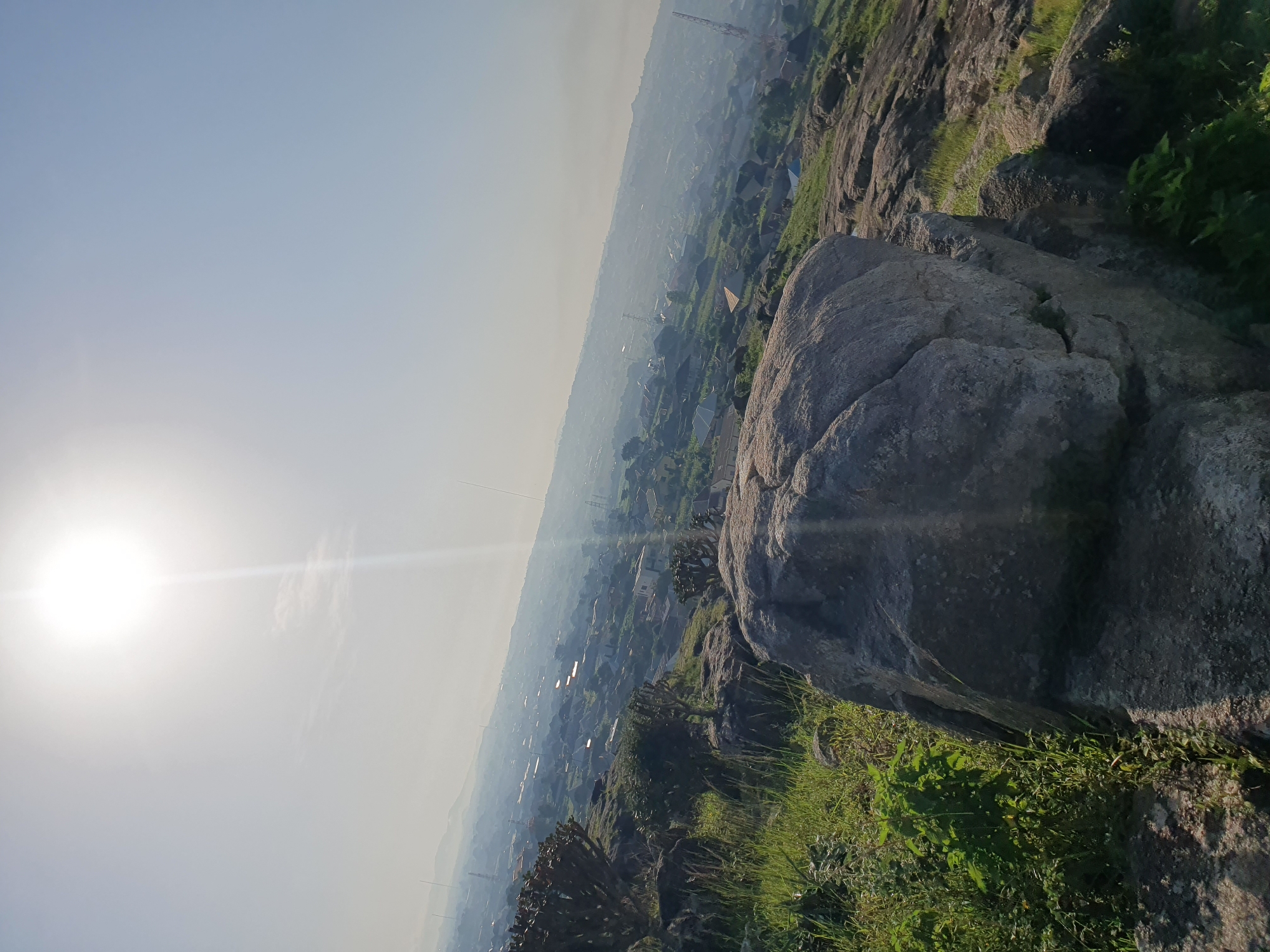 This is a high-angle, close-up, outdoor, daytime, full shot of a rocky landscape overlooking a city. The foreground is dominated by large, grey, weathered rocks with patches of green grass and vegetation. In the mid-ground, a hazy, distant city spreads out across a valley, with visible buildings, rooftops, and communication towers. The sky is bright and clear, with the sun high and slightly to the left, casting a strong light and a noticeable lens flare. The overall scene suggests a natural, elevated viewpoint above a populated urban area. The location is identified as Gura Top, Nigeria.