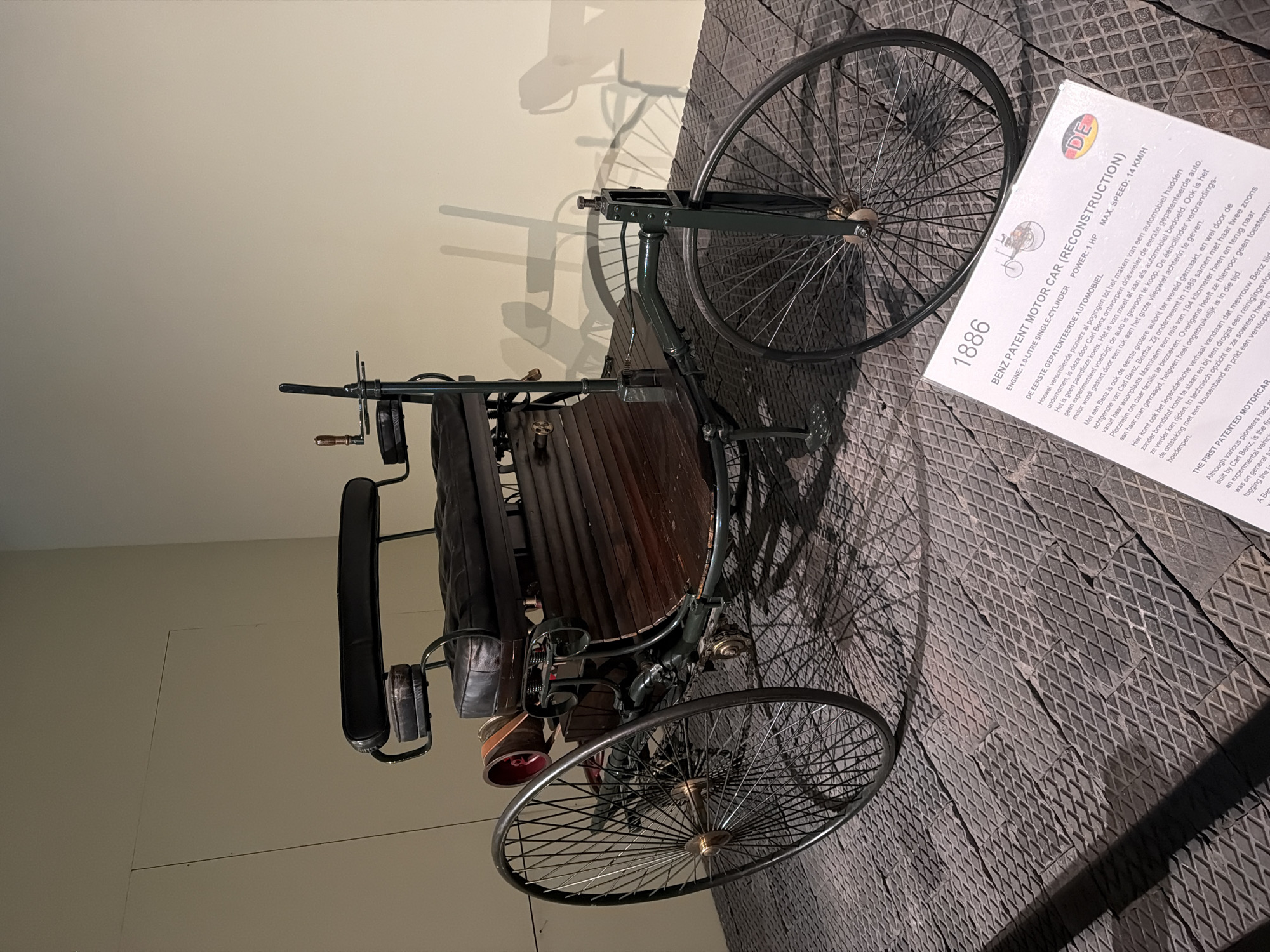 The image shows a Benz Patent Motor Car (Reconstruction) from 1886 displayed in what appears to be a museum setting. The car is a three-wheeled vehicle with a dark green frame and wooden seating. It features large spoked wheels, with the rear wheels being significantly larger than the front. A small sign in front of the car provides details about the vehicle, stating "1886 BENZ PATENT MOTOR CAR (RECONSTRUCTION) ENGINE: 1.0-LITRE SINGLE-CYLINDER POWER: 1 HP. MAX. SPEED: 14 KM/H". The sign also contains text in Dutch and English. The car is positioned on a textured floor made of dark, interlocking diamond-patterned tiles. The background is a plain, light-colored wall, with the shadow of the car cast upon it. There are no people visible in the frame. The overall impression is of a historical artifact being showcased. Based on the presence of the sign with Dutch text and the museum-like display, it is likely located in The Hague, Netherlands, possibly at a museum that features early automotive history.