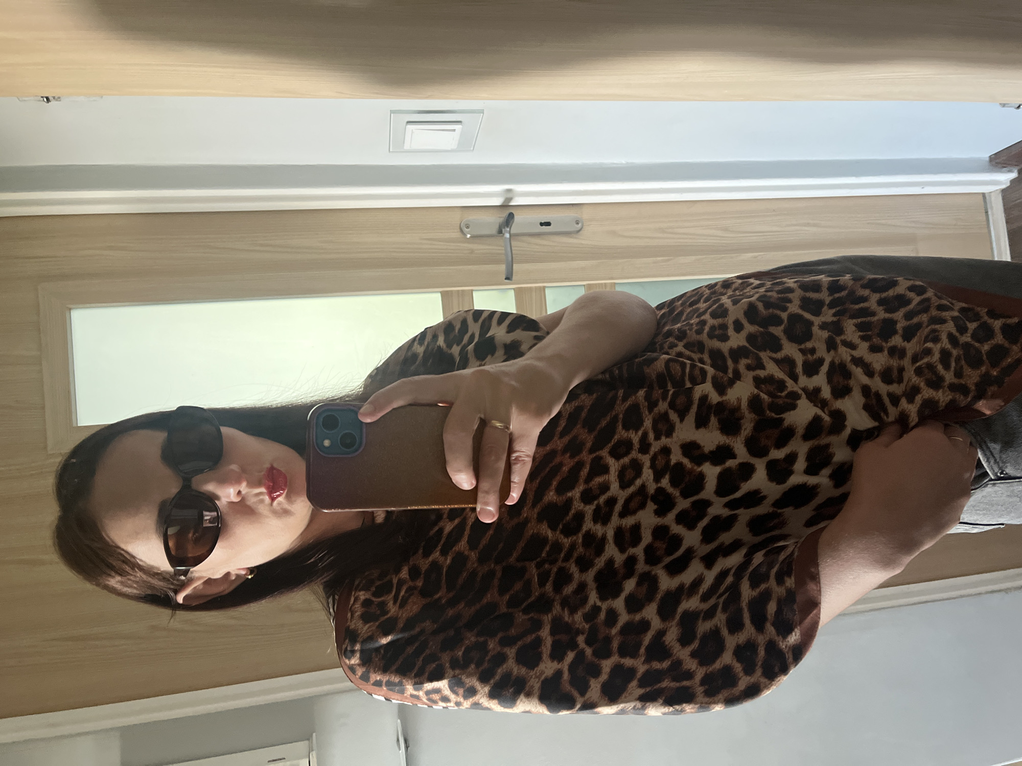 A person is captured in a mirror selfie, oriented vertically in the image. They are seen from the chest up, wearing a brown and black leopard print top or dress. Dark, cat-eye style sunglasses are perched on their face, and red lipstick is visible on their puckered lips, suggesting a playful or confident expression. Their dark hair frames their face.
The person holds a smartphone, likely an iPhone given the distinct dual camera lenses, in their right hand (as seen in the reflection). The phone has a brown or tan case. A simple gold band ring is visible on their ring finger of the hand holding the phone.
The background reveals an indoor setting, dominated by light-colored wooden paneling or a door. A silver-toned handle or latch mechanism is affixed to the wooden surface behind their right arm. Above this, a white wall section features white molding and a rectangular recessed ceiling light fixture. The overall lighting appears bright, suggesting a well-lit interior, possibly during the daytime. No visible text is present in the image.