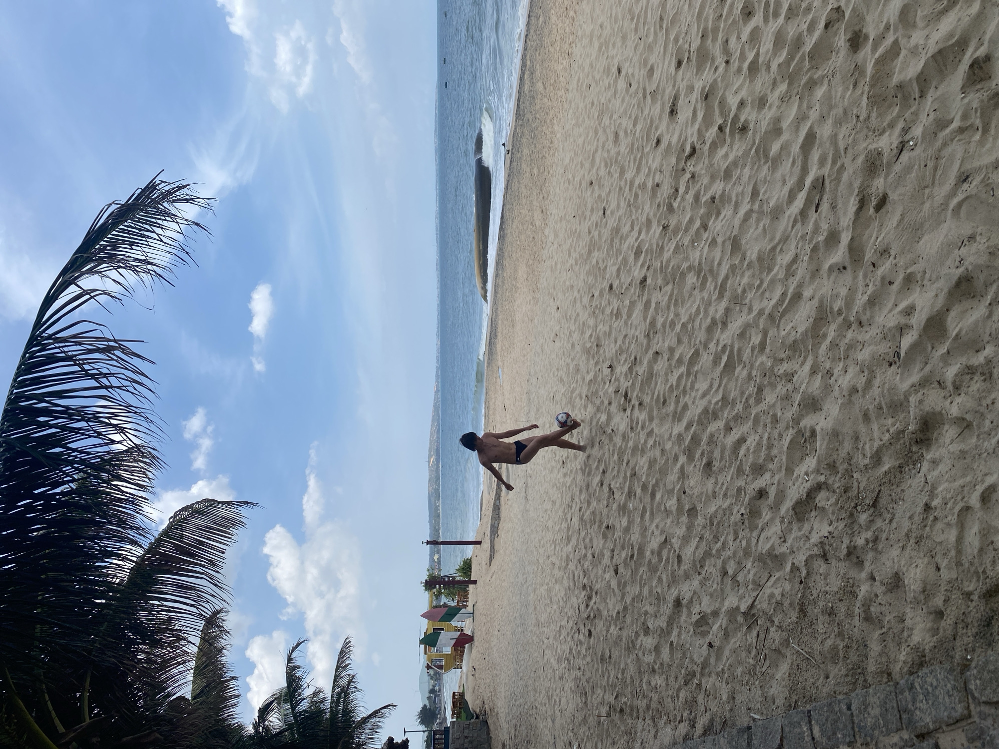 The image depicts a person playing soccer on a sandy beach. The person, wearing black swim trunks, is captured in mid-air, kicking a soccer ball with their right foot. The background shows the ocean with gentle waves and a clear blue sky with scattered clouds. Palm tree fronds are visible in the foreground on the left side of the image. In the distance, there are some beach umbrellas and structures. The lighting suggests it is daytime, likely in the late morning or afternoon, with bright sunlight illuminating the scene. The sand is textured with footprints and ripples, indicating recent activity. The overall atmosphere is one of recreation and leisure on a tropical beach.